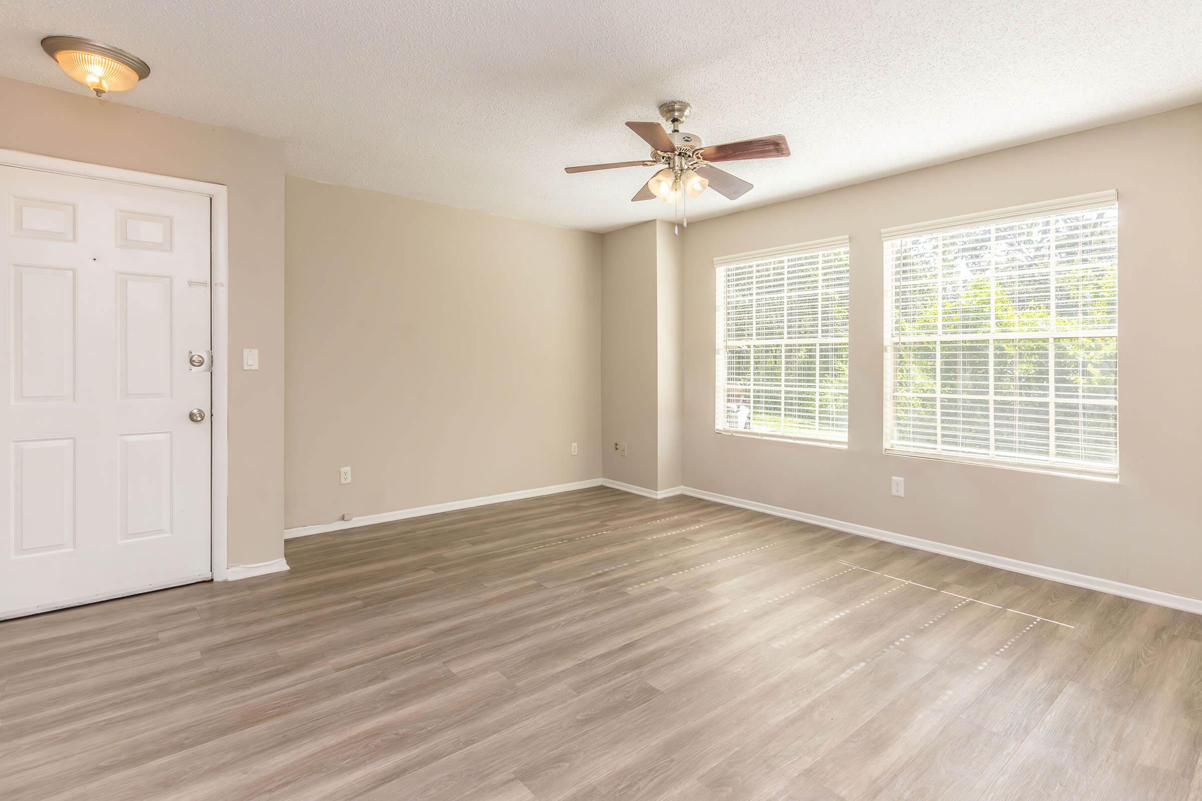 A spacious, empty room with light-colored walls and a ceiling fan. The floor features light wood-like laminate, and there are large windows with white blinds allowing natural light in. The room includes a front door with a light fixture above, creating a welcoming entrance.