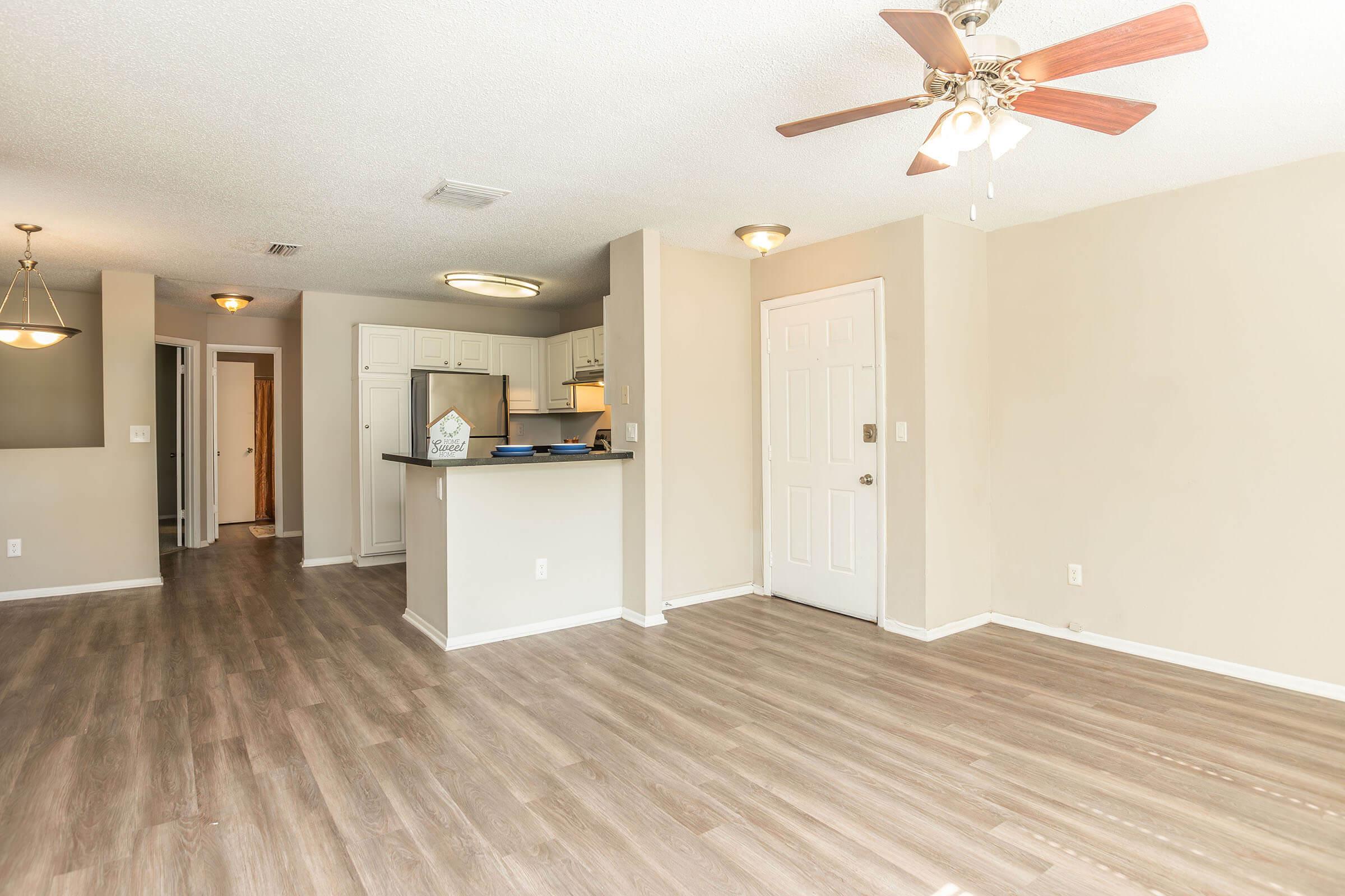 Interior view of a light-colored, spacious living area featuring a ceiling fan, wood-like flooring, and a kitchen area partially visible. There are doorways leading to other rooms, and the walls are painted in soft neutral tones, creating a bright and inviting atmosphere.