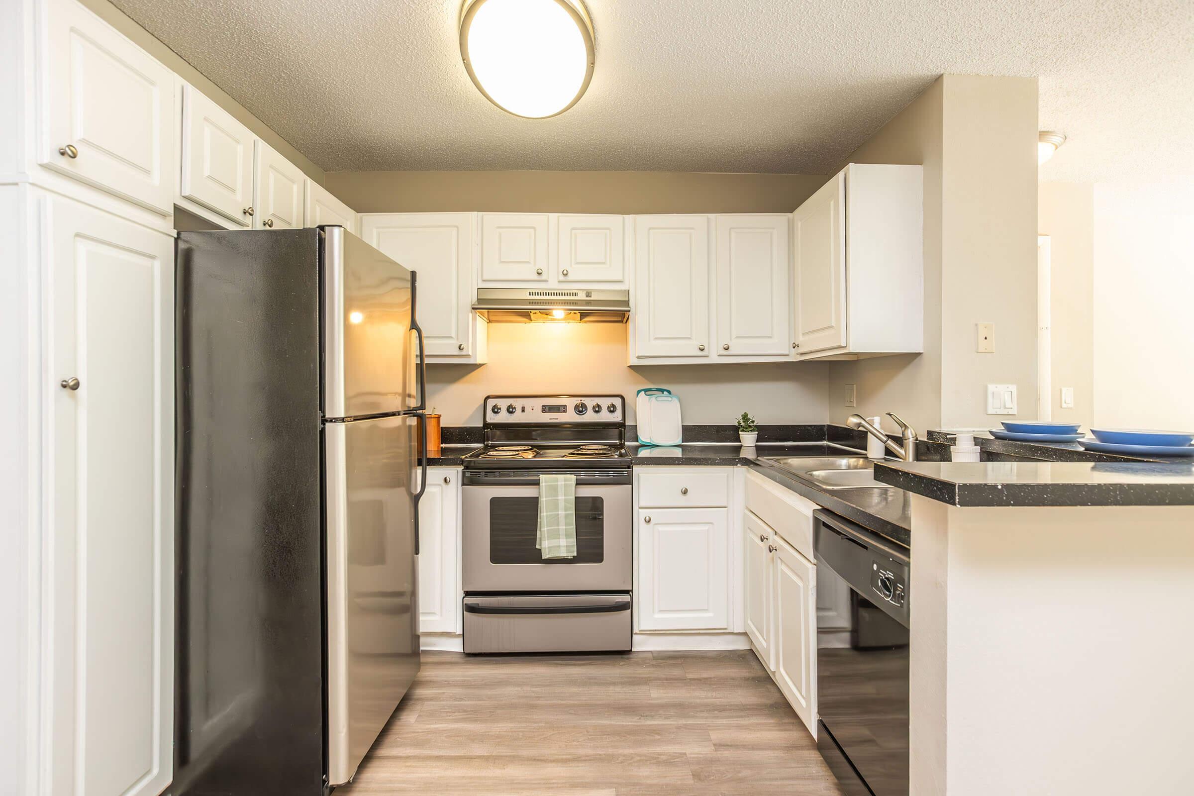 A modern kitchen featuring white cabinets, a stainless steel fridge, an oven, and a dark countertop. The floor is a light wood finish, and there are small decorative items on the counter. The space is well-lit with a ceiling light and has a clean, organized appearance.