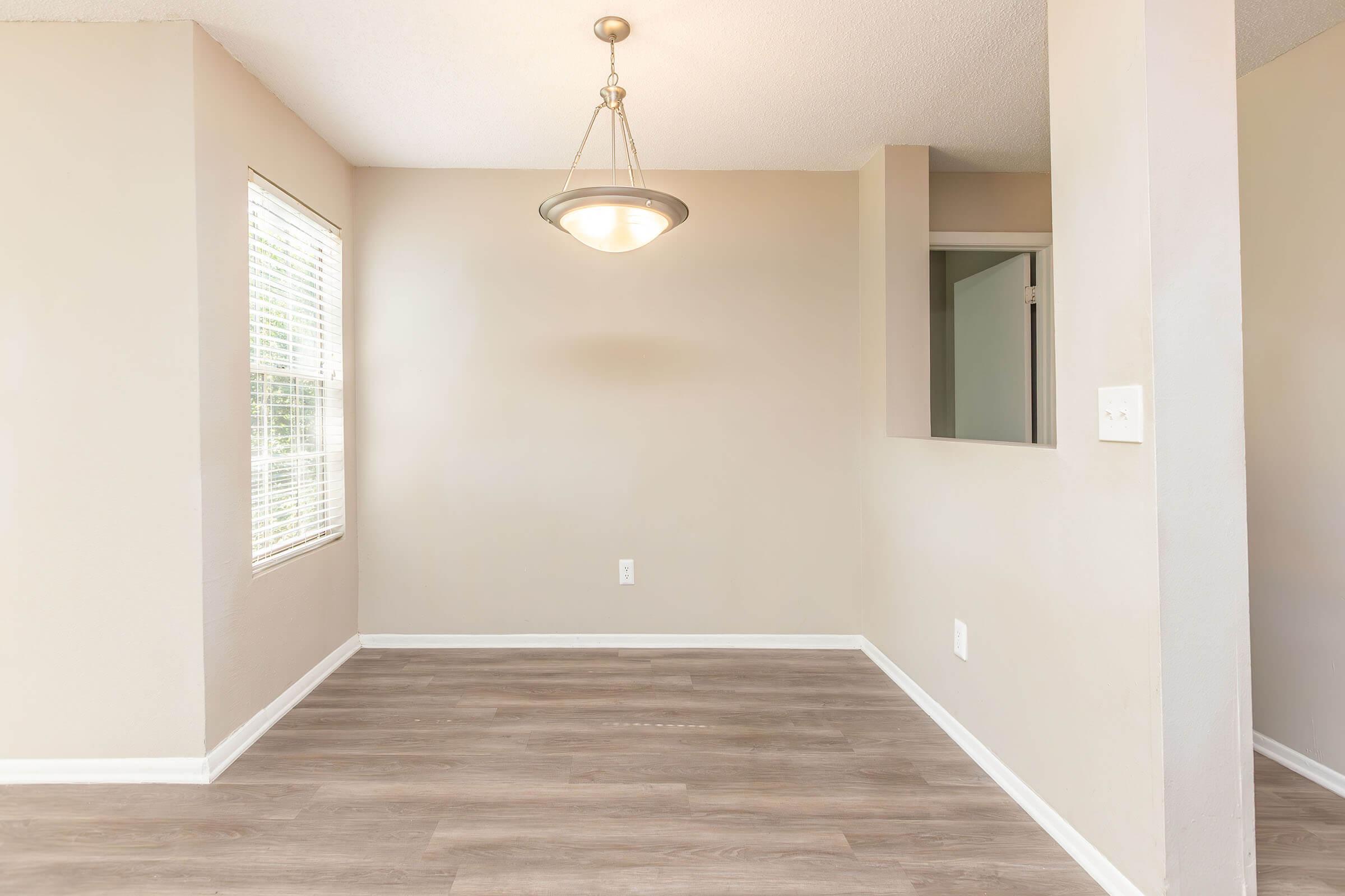 A well-lit room with a beige wall and wooden flooring. A ceiling light is hanging above, and there is a window on the left with blinds. To the right, a small opening leads to another space, possibly a kitchen. The overall atmosphere is clean and inviting, suitable for dining or relaxation.