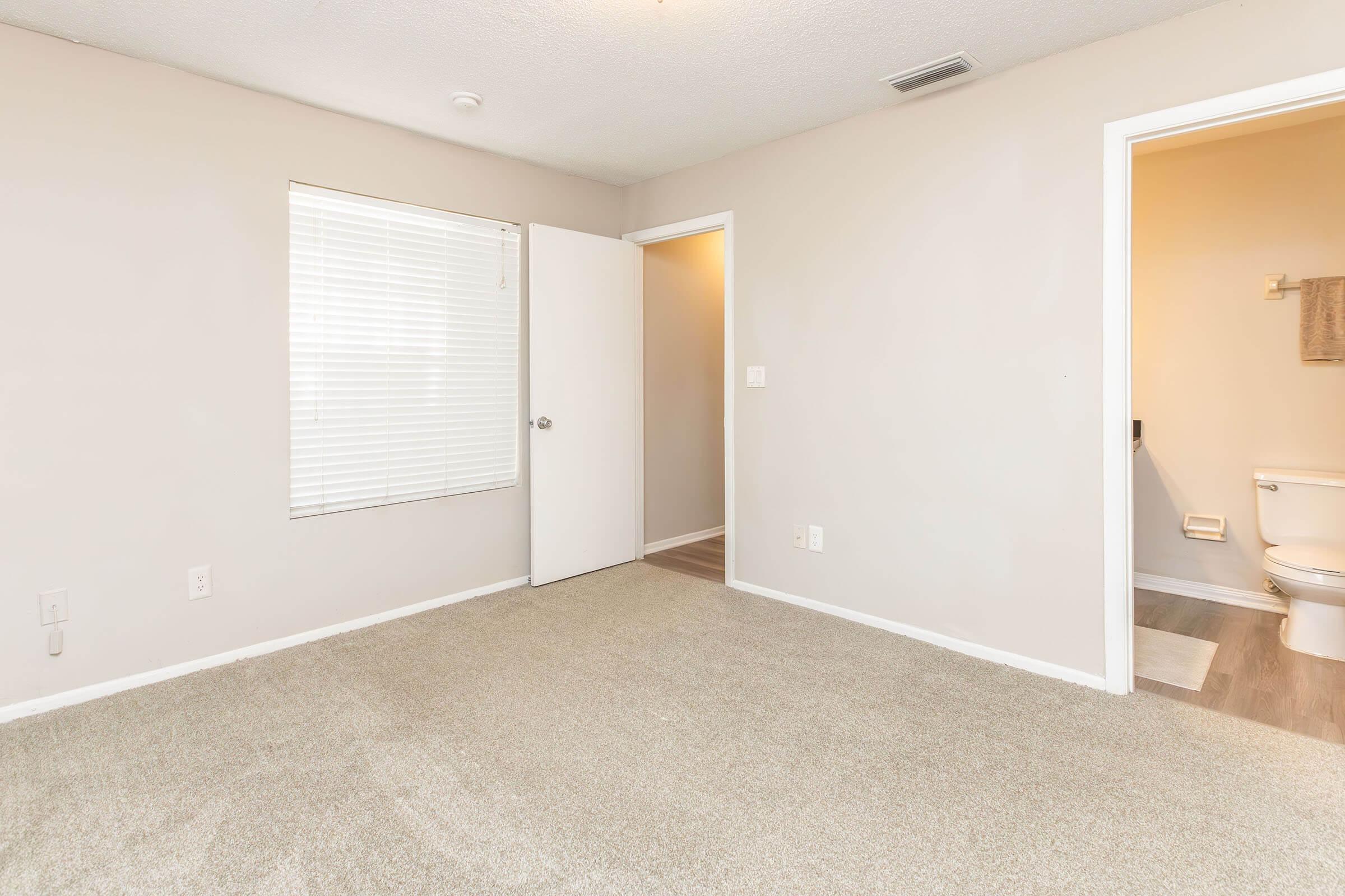 A light-colored, empty room with beige carpet and plain walls. There is a window covered by blinds on the left side, a doorway leading to another room on the left, and a door to a bathroom visible on the right, showcasing a toilet within the bathroom. The overall atmosphere is clean and simple.