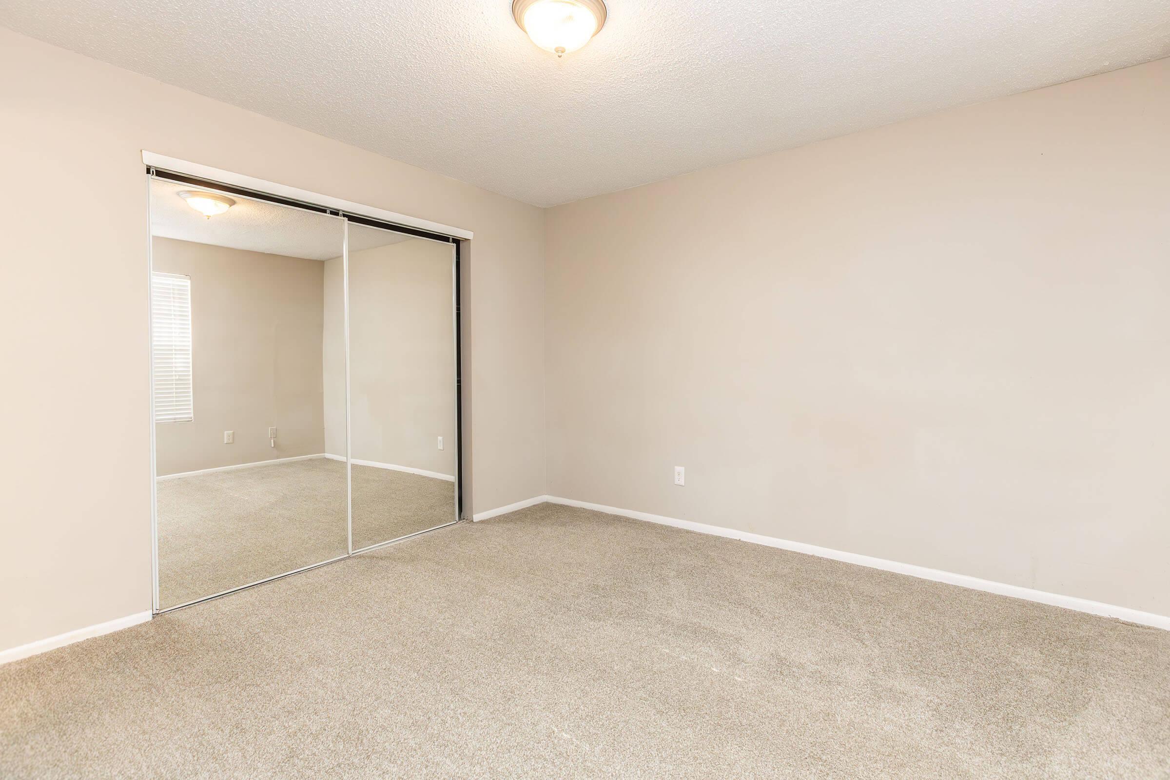 Empty room with beige walls and carpeted floor. A large mirrored closet reflects light, and a ceiling lamp provides illumination. A window allows natural light to enter, casting soft shadows on the floor. The space appears clean, ready for furnishing or decoration.