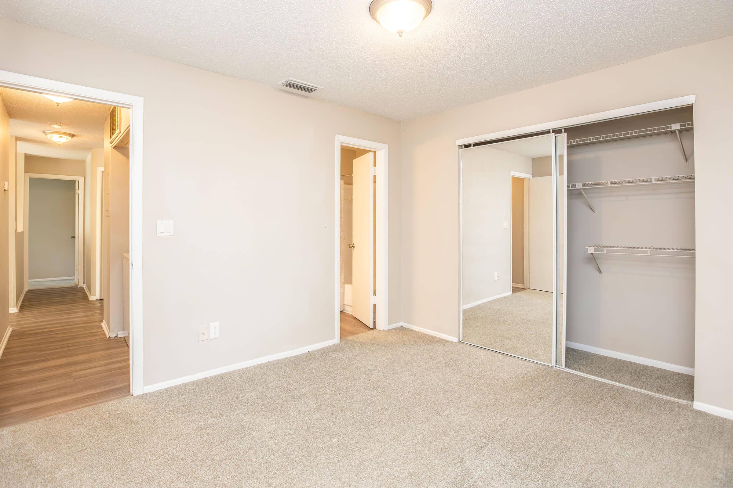 A spacious, empty bedroom with light-colored walls and carpet. There's a mirrored closet with shelving and two doorways leading to other rooms. The lighting is bright from a ceiling fixture, creating an open and inviting atmosphere.