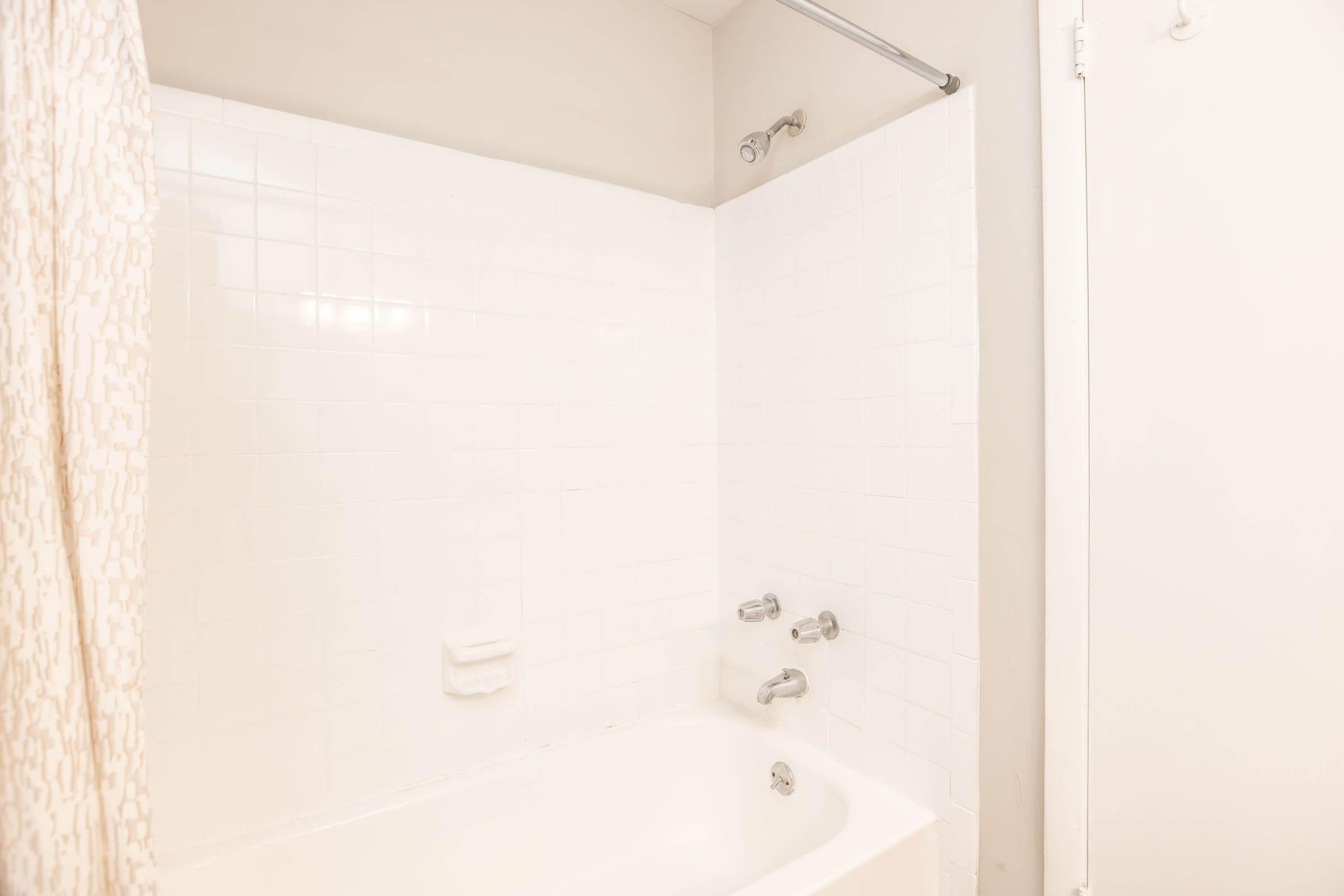 A clean and simple bathroom with a white tiled shower area, a bathtub, a shower curtain, and chrome faucets. The walls are light-colored, and there is a towel bar mounted on the wall.