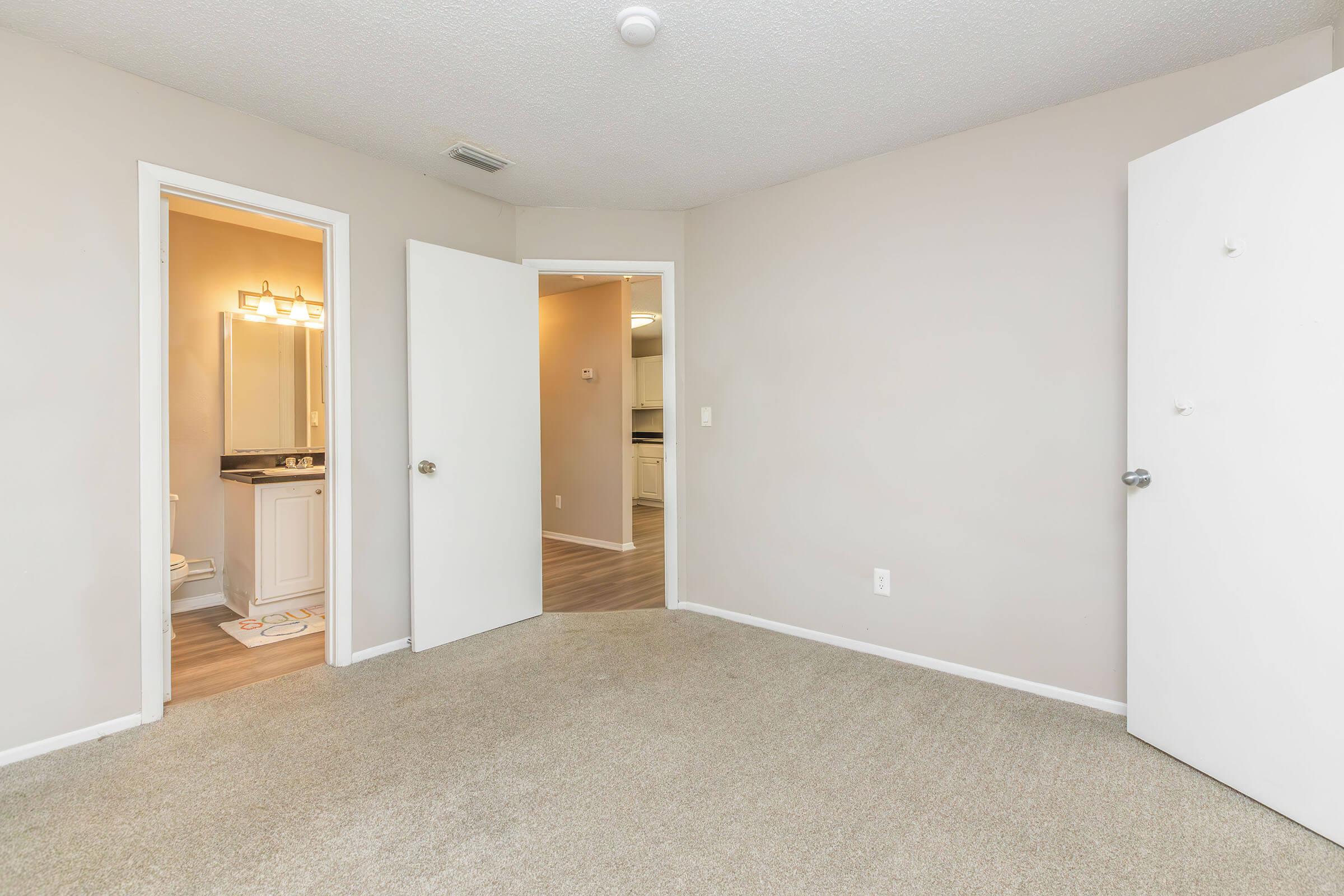 A view of a room featuring light-colored walls and beige carpet. Two open doors lead to a bathroom on the left with a mirror and a vanity, and a kitchen area on the right. The room has no furnishings, creating an empty, spacious appearance.