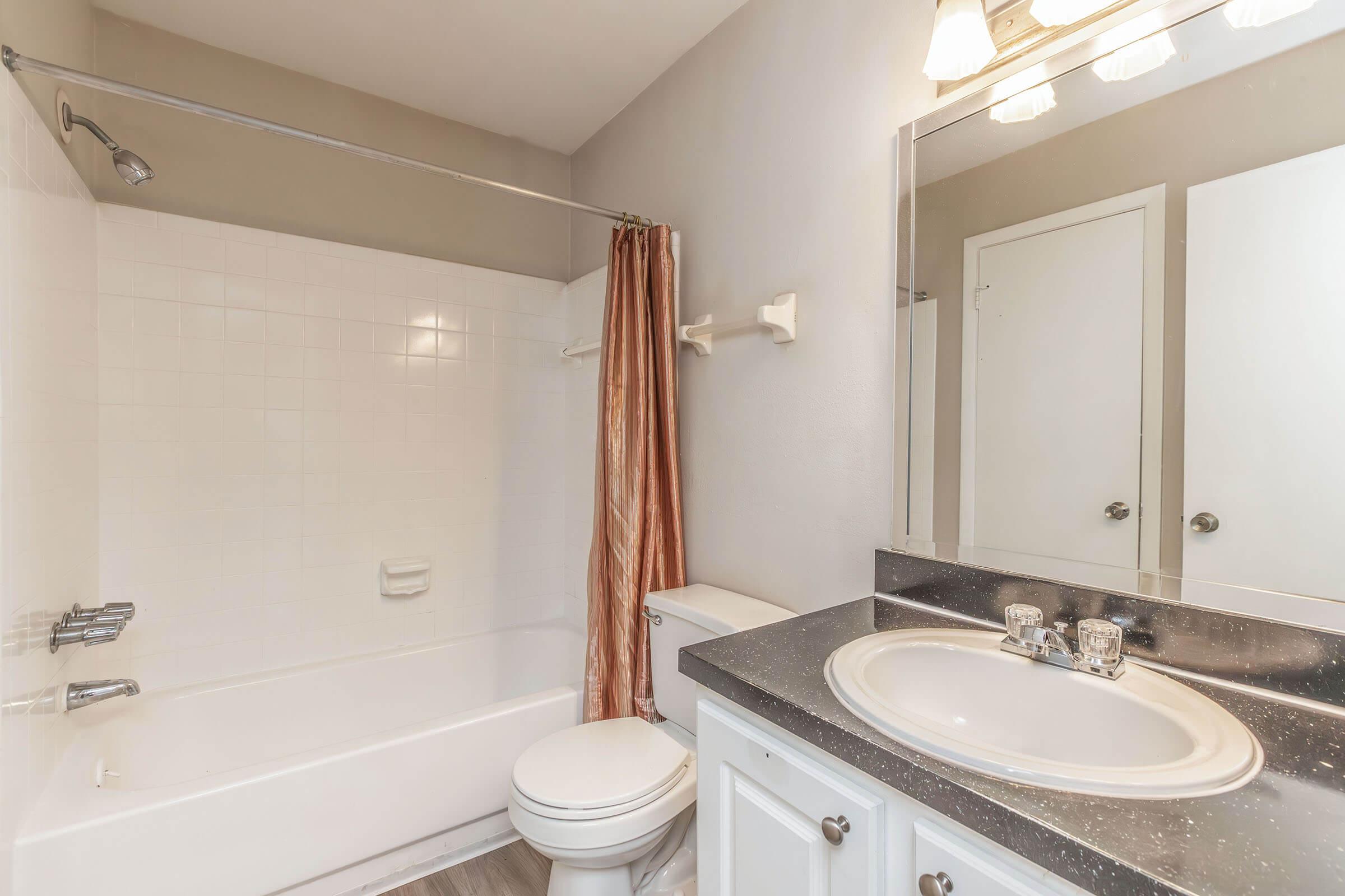 A well-lit bathroom featuring a white bathtub with a shower curtain, a toilet, and a sink with a dark countertop. The walls are painted in a neutral color, and there is a large mirror above the sink with three light fixtures. The floor has a simple finish, creating a clean and modern look.