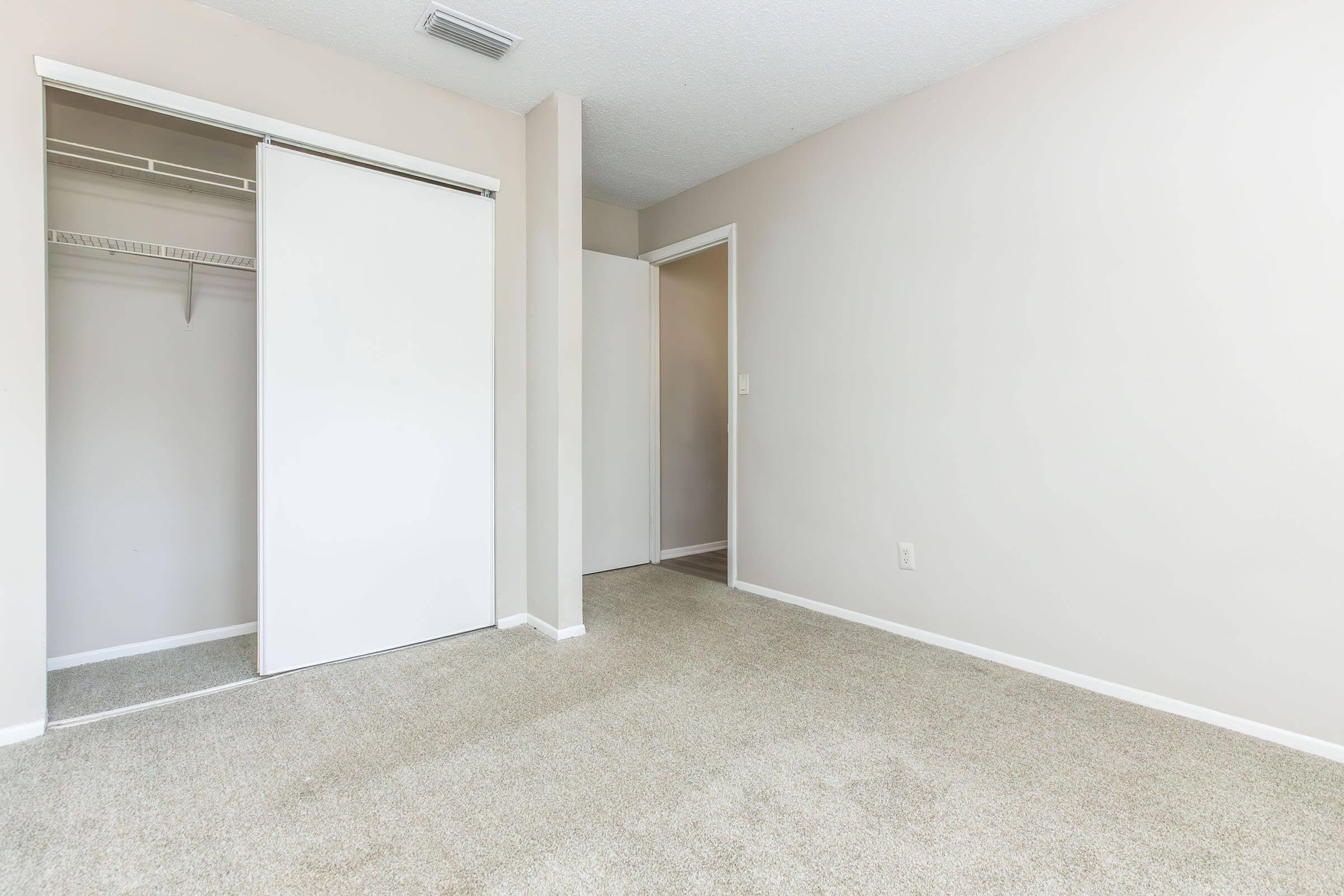 A vacant room with light-colored walls and beige carpet. There is a closet with sliding doors on the left and an open doorway leading to another area on the right. The space is well-lit and empty, creating a clean and simple atmosphere.