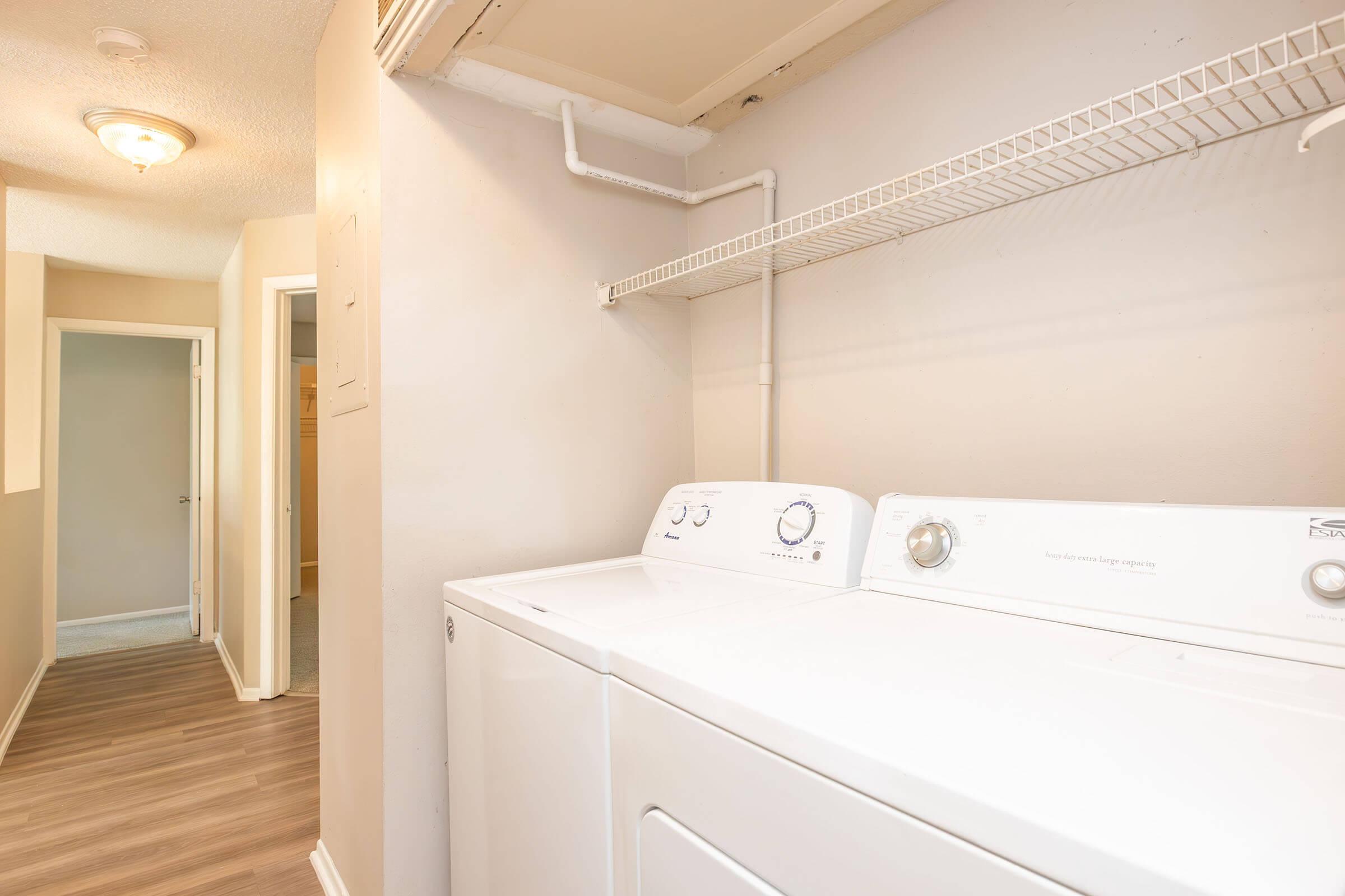 Laundry room featuring a washing machine and dryer with a shelf above them. The walls are light-colored, and there is a doorway leading to another room in the background. The flooring is a light wood look. Natural light is coming from a ceiling fixture.