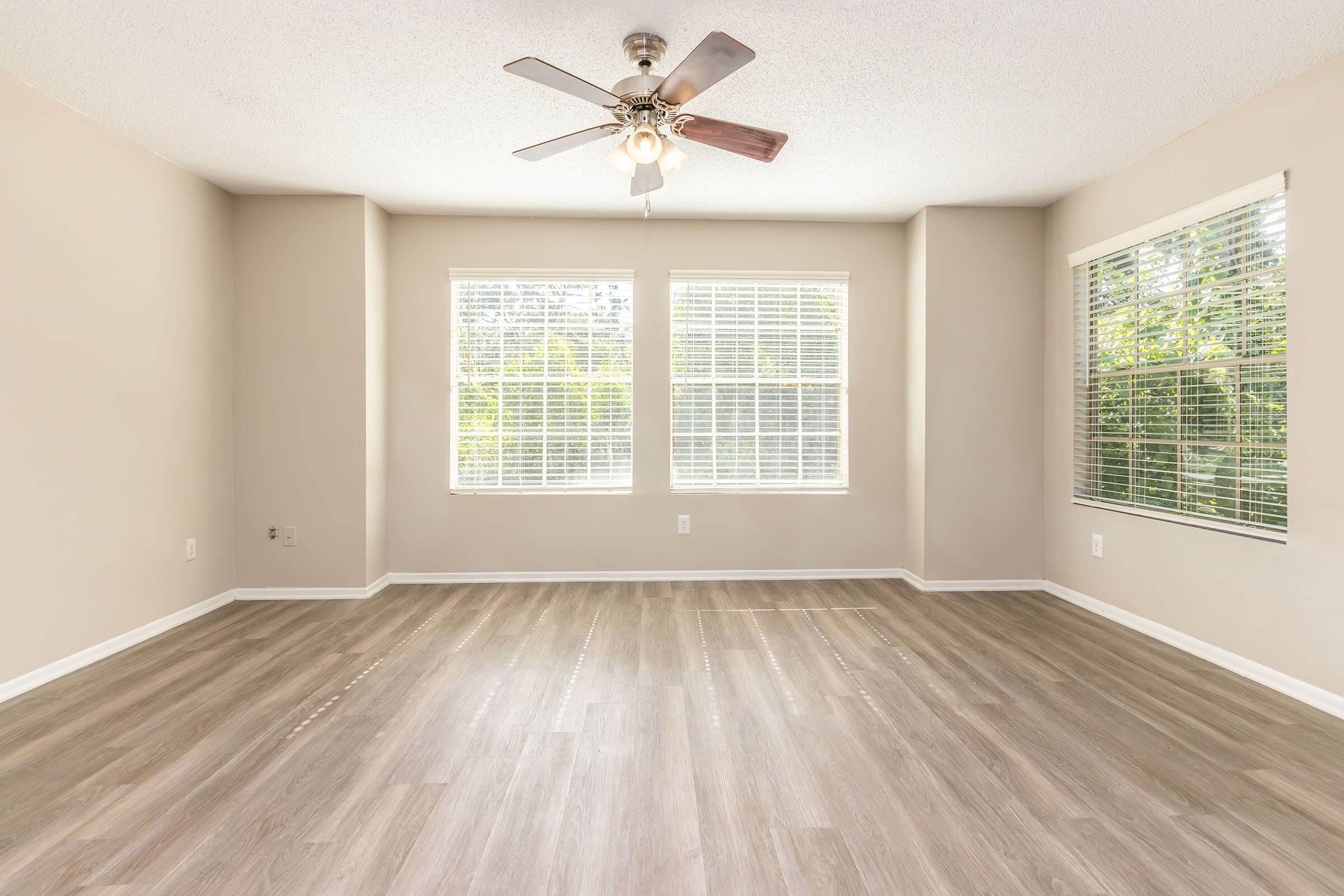 A spacious, well-lit room featuring a ceiling fan, large windows with white blinds, and light-colored walls. The floor is finished with a modern, light wood-like finish, creating a bright and inviting atmosphere.