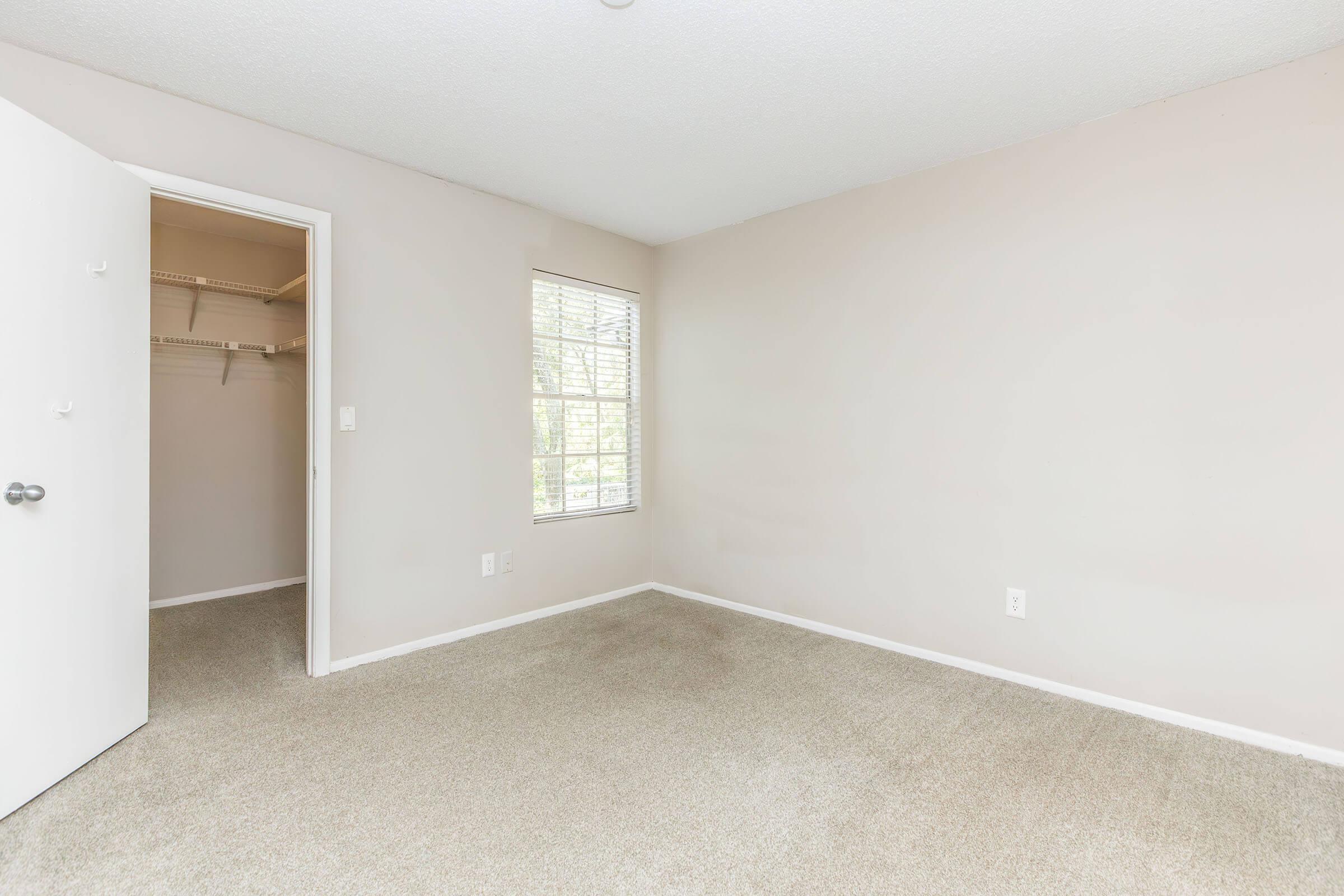 A clean, empty bedroom featuring light beige walls and carpet. There is a small window allowing natural light, and a closet with sliding doors on one side. The room has a simple, uncluttered appearance, making it ideal for personalization and decoration.