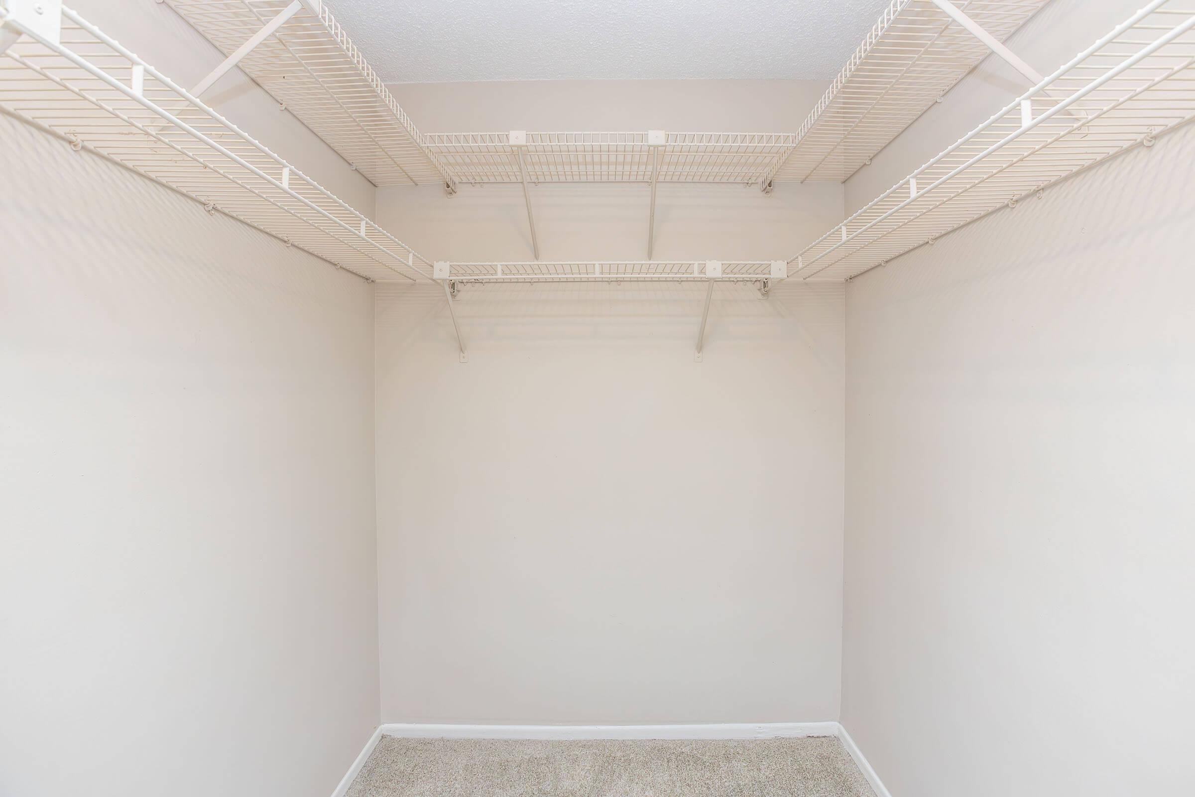 Empty closet space with white wire shelving mounted on the walls. The walls are painted light beige, and the floor is carpeted in a neutral color. There are no items visible in the closet, creating a clean and open storage area.