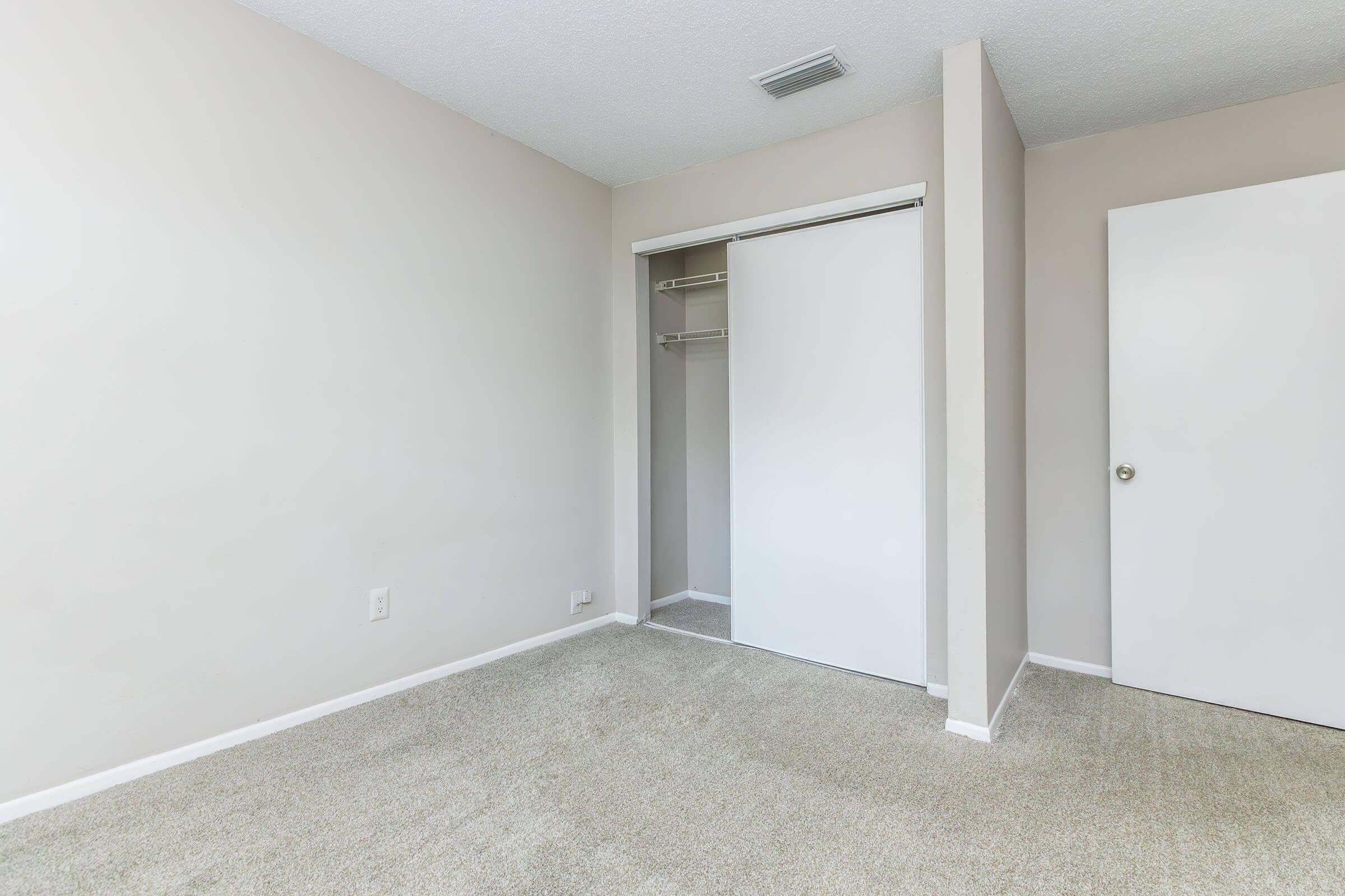 A spacious, empty bedroom with light beige walls and soft carpet. The room features a white sliding closet door and an adjacent closed door, creating a clean and simple aesthetic. Natural light is visible, enhancing the airy atmosphere of the space.