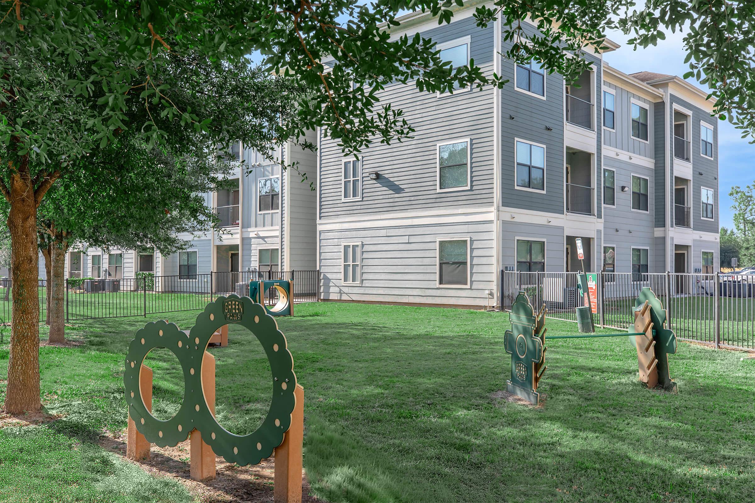 Landscaped area with green grass and decorative sculptures. In the foreground, two framed shapes with cutouts and a textured sculpture are visible. In the background, a multi-story apartment building features gray siding and several windows, surrounded by trees and a fence.