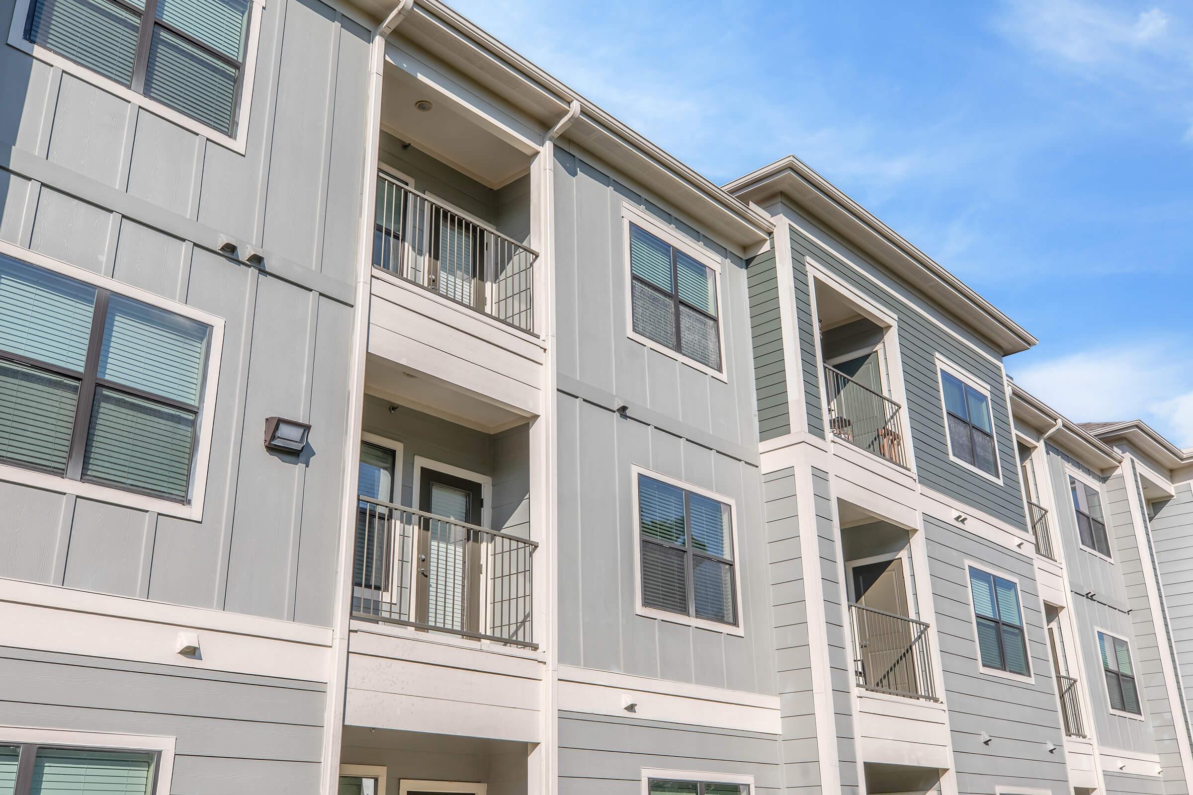 Modern apartment building exterior featuring multiple stories, with balconies and large windows. The facade is a mix of gray and green siding under a bright blue sky, showcasing a contemporary architectural design.