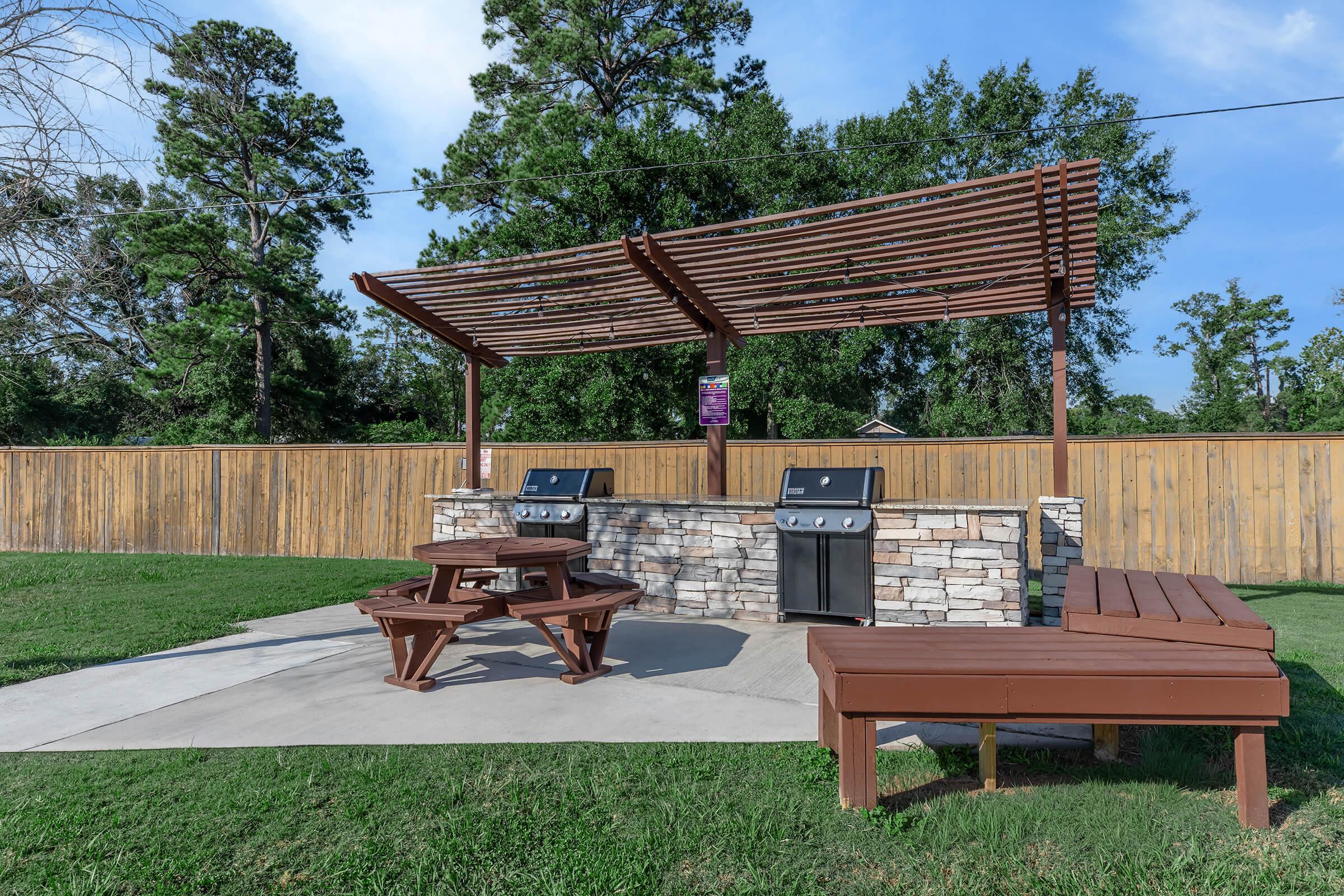 An outdoor grilling area with two barbecue grills under a wooden pergola, surrounded by a grassy lawn. There are wooden picnic tables for seating, and a wooden fence in the background provides privacy. The sky is clear with a few clouds, suggesting a pleasant day.