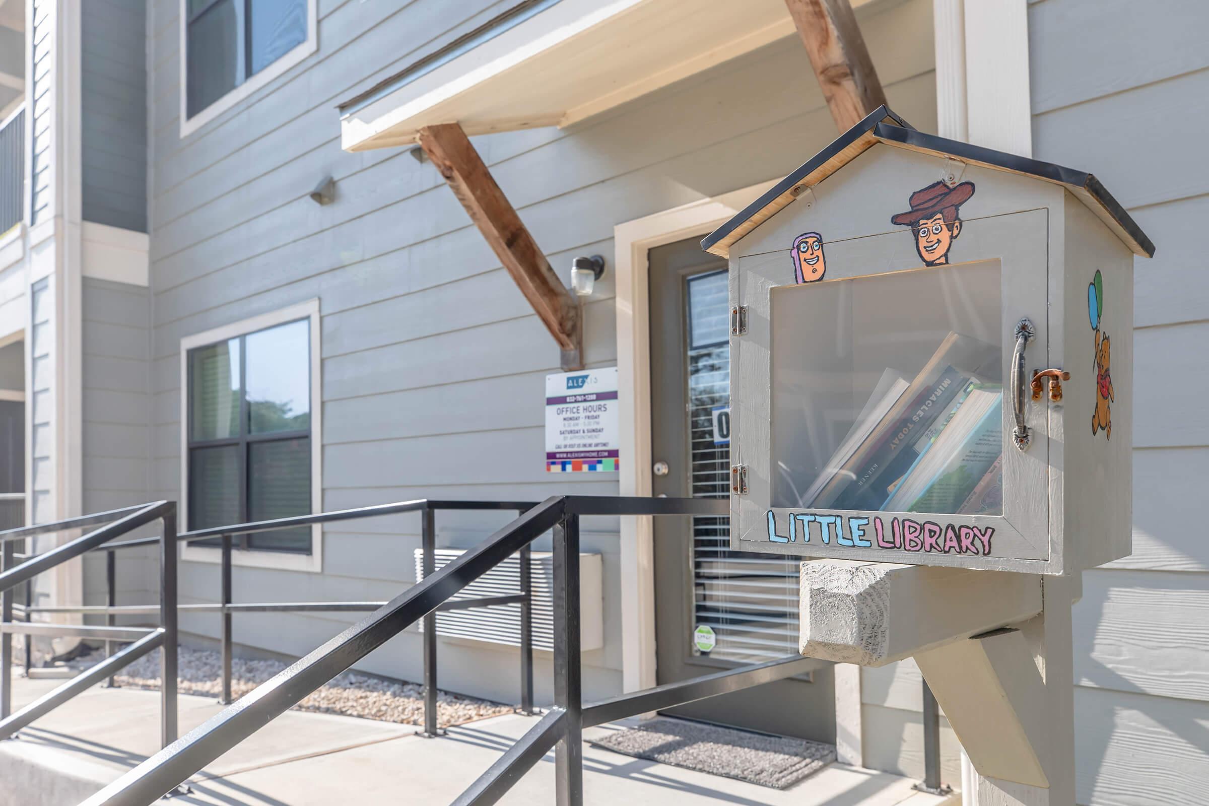 A small, charming "Little Library" box mounted on a post, filled with books, featuring playful artwork of characters on its front. It is situated outside a modern building with gray siding and large windows. A sign is visible nearby, indicating the location of the library.
