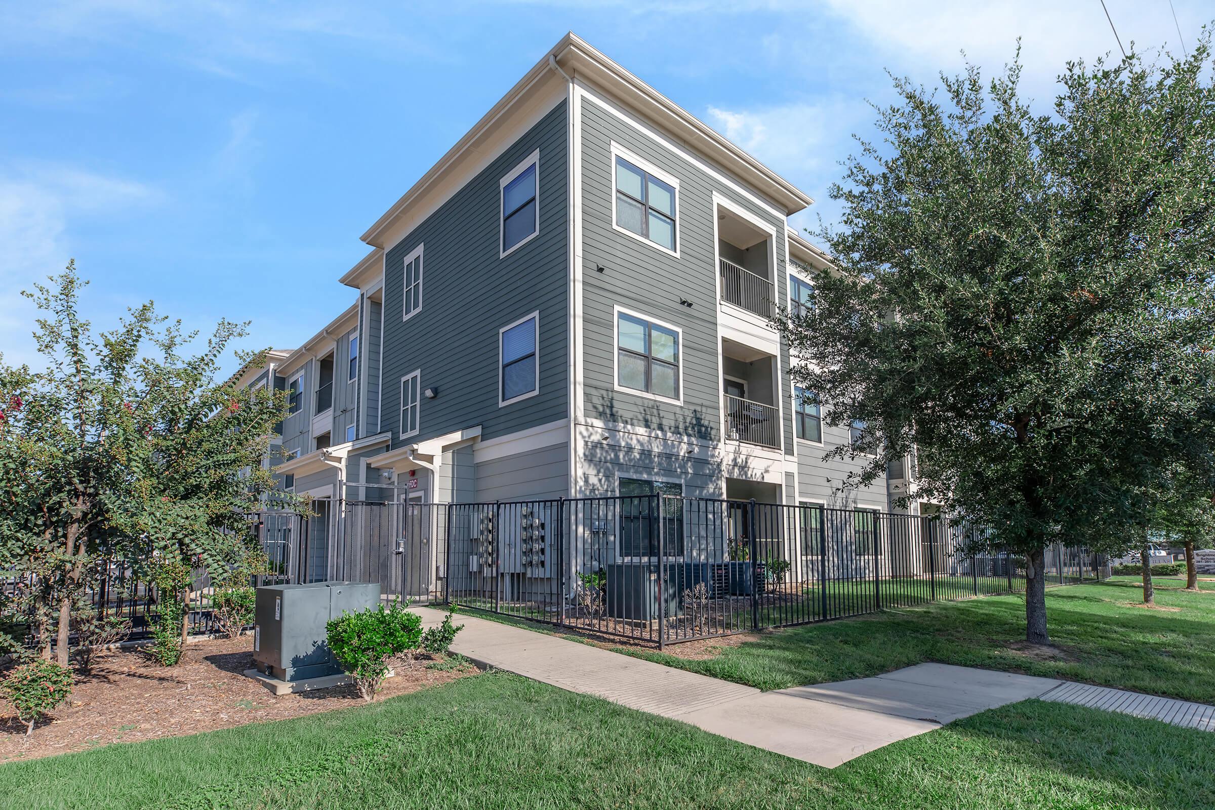 Exterior view of a modern three-story apartment building with gray siding. The building features balconies and a landscaped area with grass, shrubs, and trees. A concrete pathway leads up to the entrance, and there is a gated fence surrounding the property. Clear blue sky in the background.