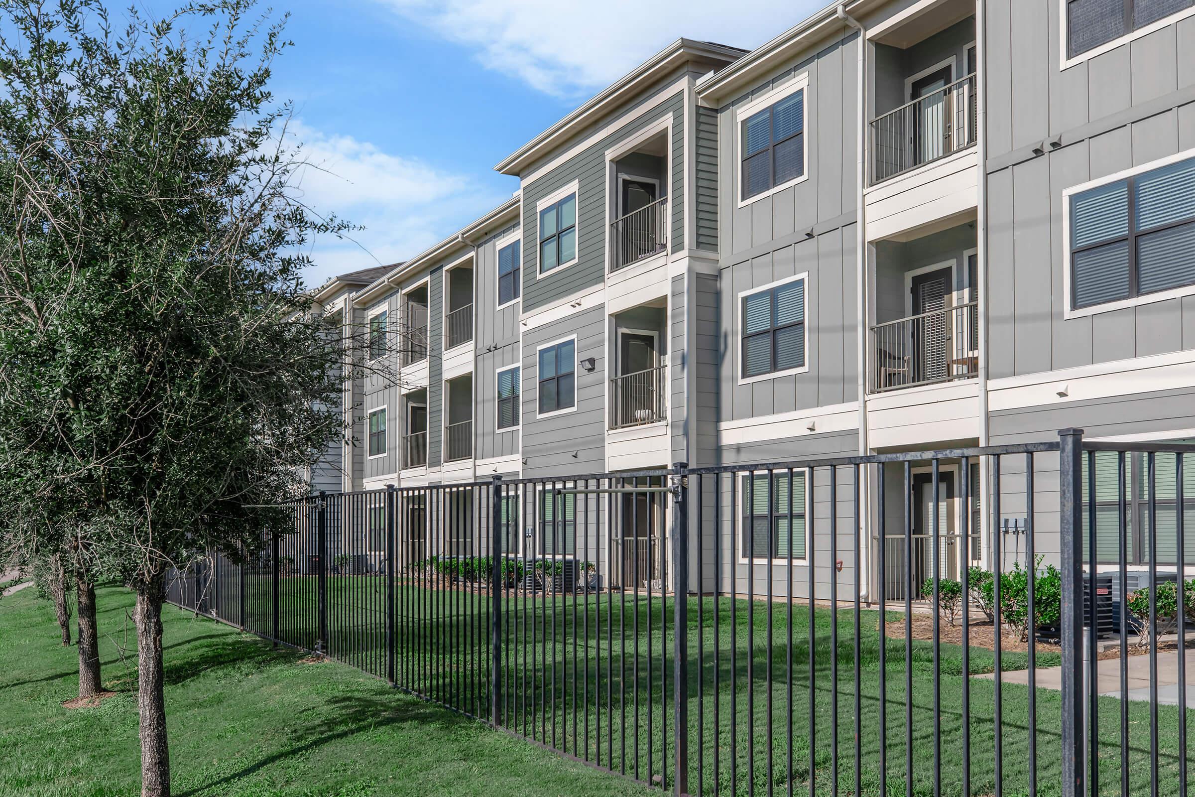 A row of modern apartment buildings with multiple stories, featuring balconies and green grass in front. A black metal fence runs along the perimeter, and there are small trees and shrubs in the landscaping. The sky is partly cloudy with blue visible.