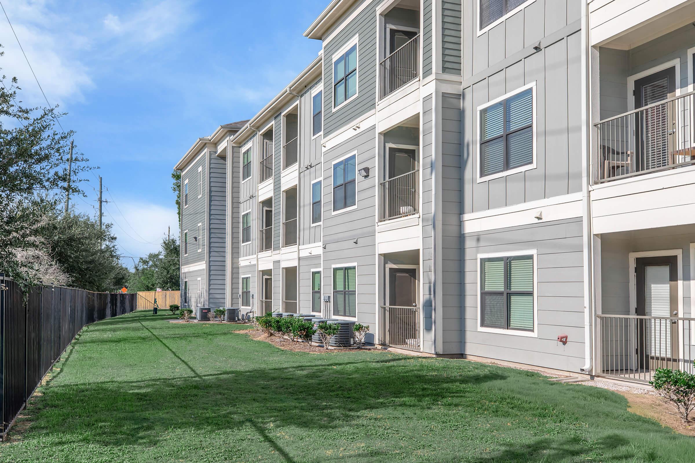 View of a modern apartment complex featuring multiple stories with light gray siding, balconies, and large windows. A well-maintained lawn with green grass is visible in front, alongside a fenced area and a clear blue sky in the background.