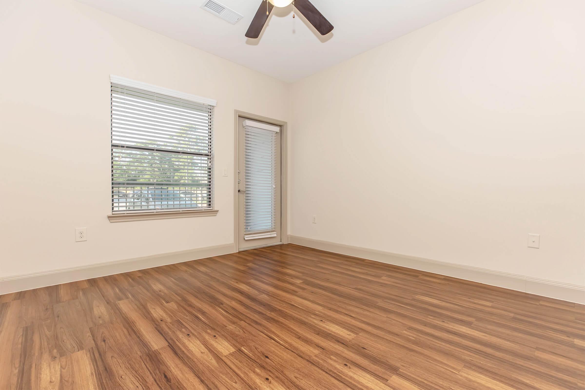 Empty room with light-colored walls and a ceiling fan. The floor features dark wood laminate. A window with white blinds lets in natural light, and there is a sliding glass door leading outside. The space is clean and unfurnished, highlighting the flooring and natural light.