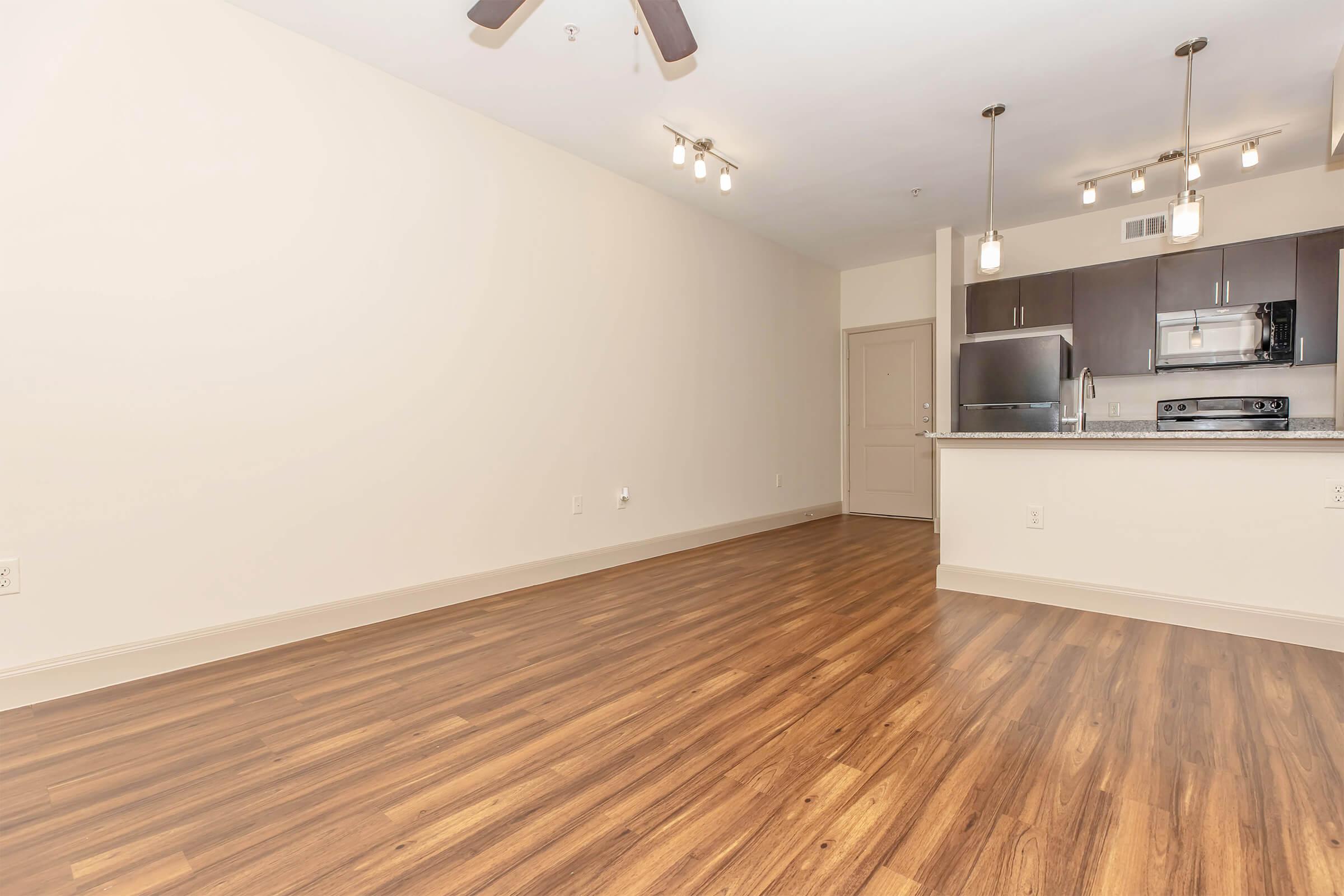 Interior view of a modern apartment featuring a spacious living area with hardwood flooring, a ceiling fan, and an open kitchen in the background. The kitchen includes stainless steel appliances and pendant lighting. The walls are light-colored, creating a bright and airy atmosphere.