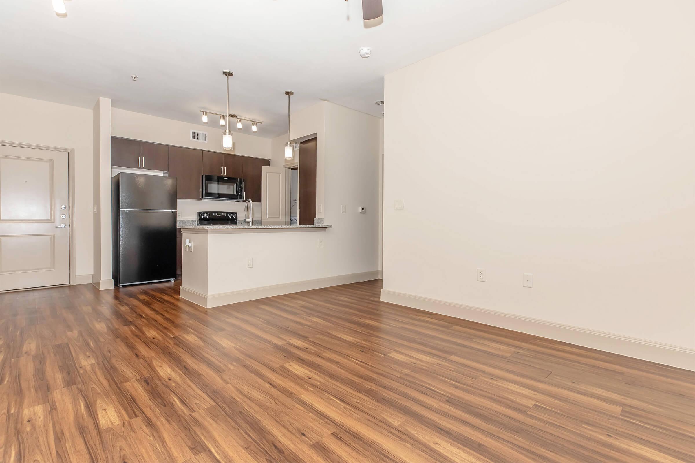 Modern apartment interior featuring hardwood floors, a spacious living area, and an open kitchen with stainless steel appliances. The kitchen includes a refrigerator, microwave, and dark cabinetry. Soft lighting fixtures hang from the ceiling, enhancing the bright, airy atmosphere.