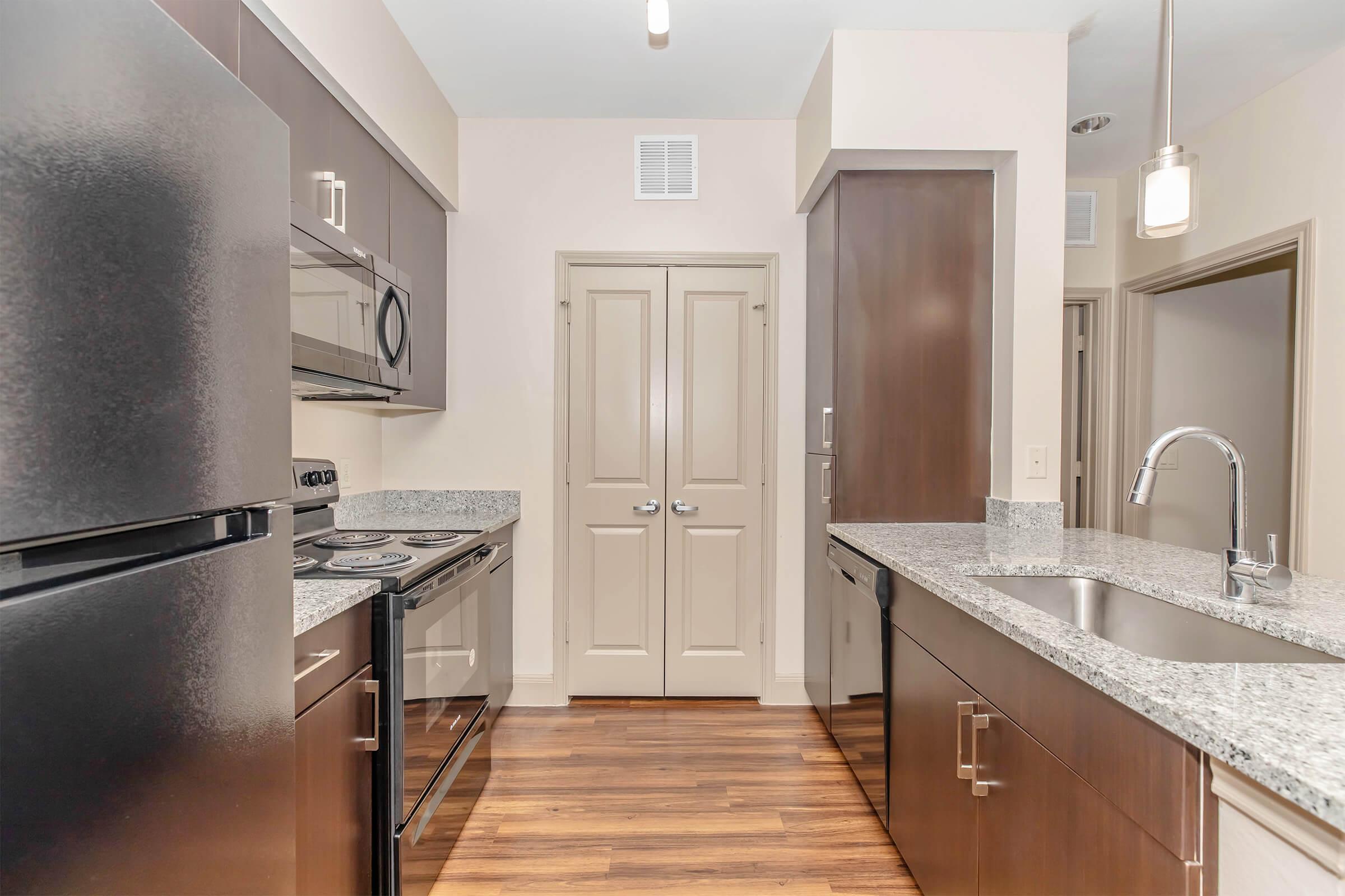 Modern kitchen featuring dark wood cabinetry, granite countertops, stainless steel appliances, and a sleek black refrigerator. Includes an oven, microwave, and sink with a modern faucet. The space has wood-style flooring and a door leading to another room. Bright lighting enhances the contemporary design.
