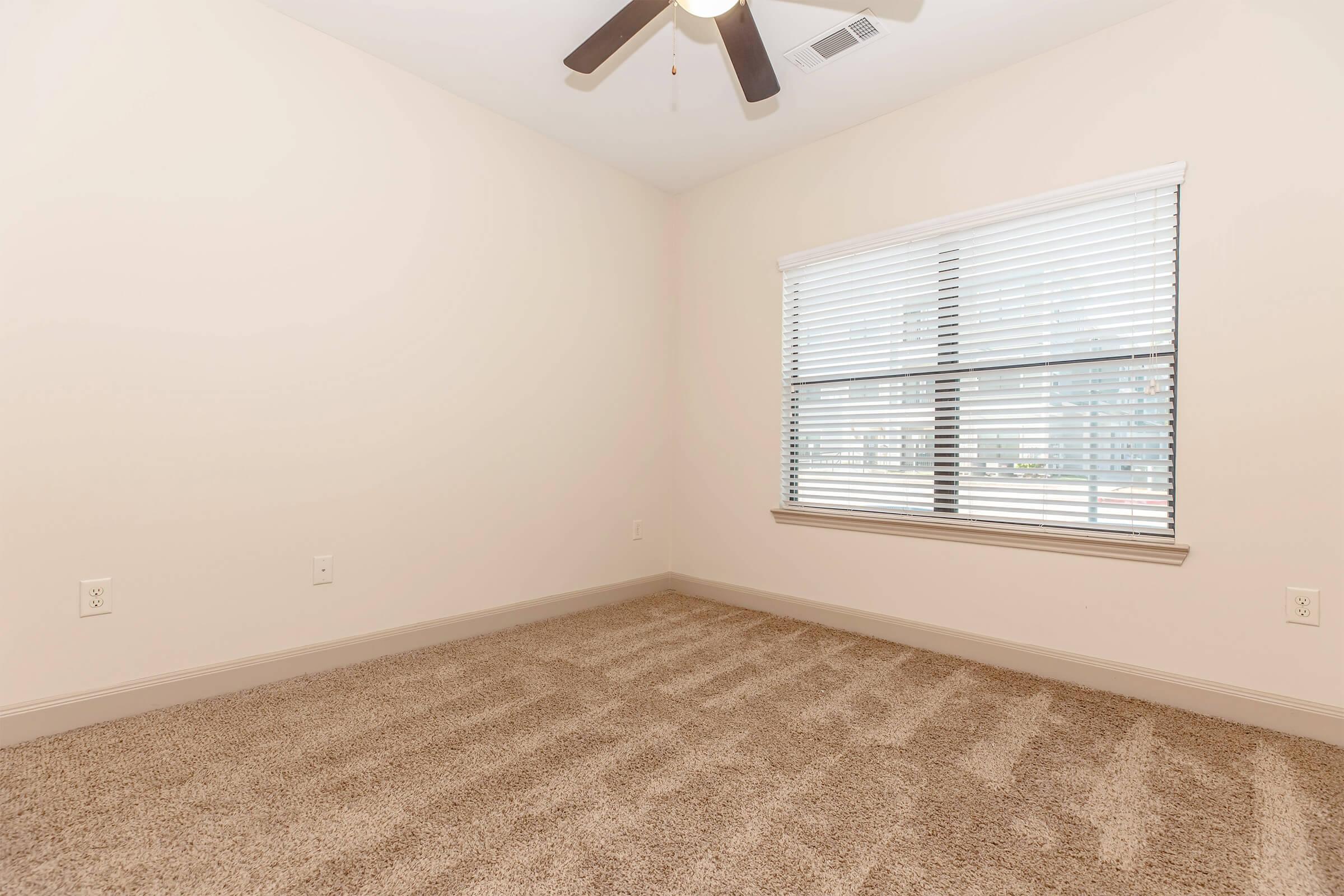 Empty bedroom with light beige walls and a window covered by blinds. The carpet is soft and tan-colored, and a ceiling fan is visible. The room is bright and spacious, suggesting a neutral and inviting atmosphere.