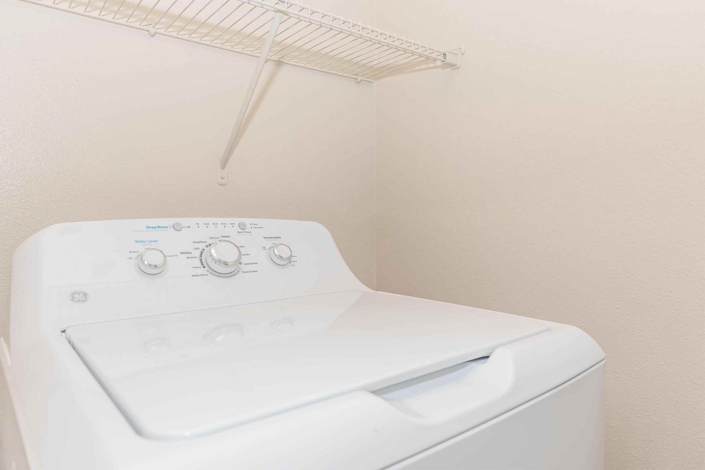 A white washing machine with control dials and settings, placed in a light-colored laundry room with a plain wall and a wire shelf above it. The machine is clean and appears to be in good condition.