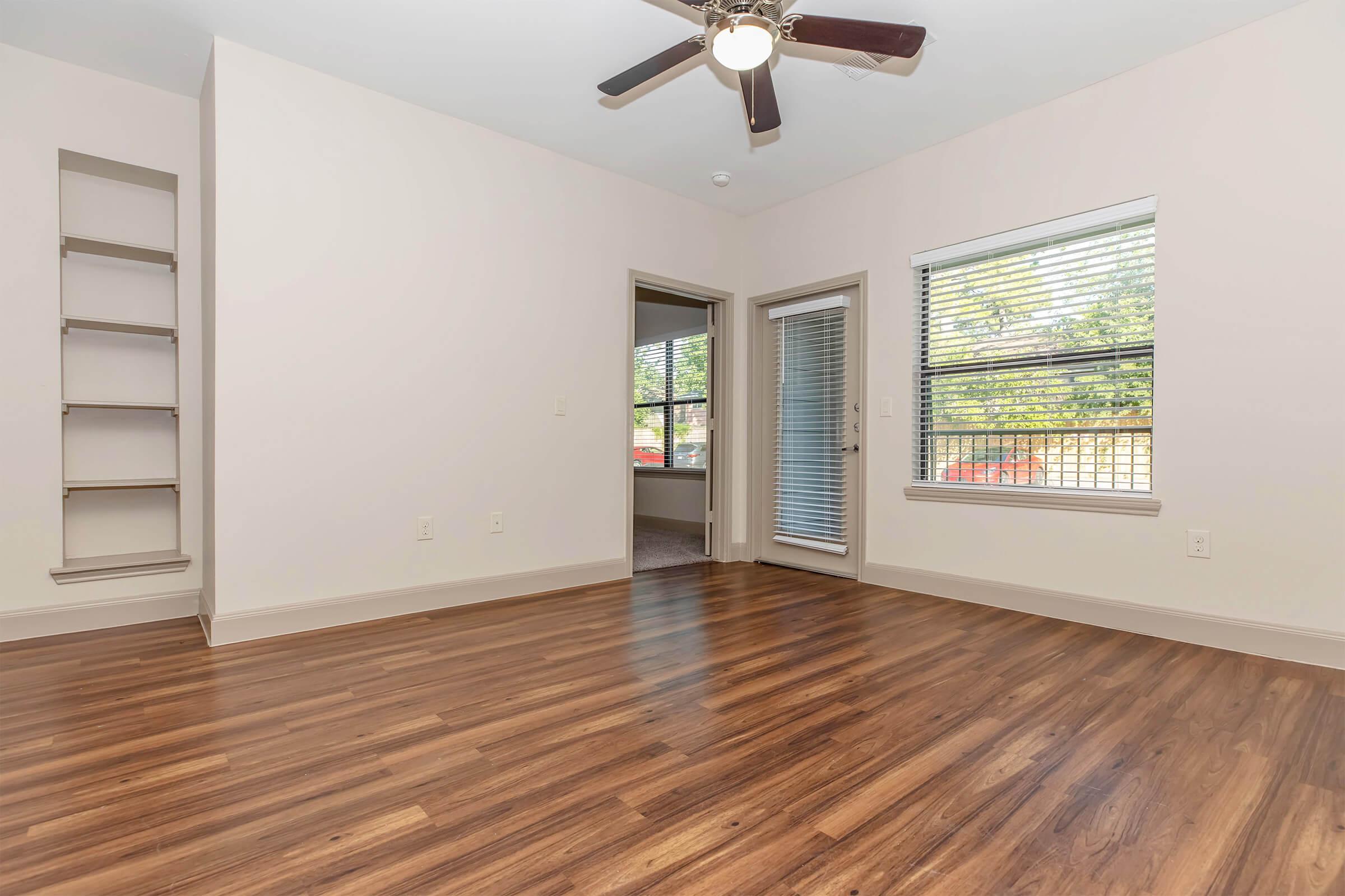 A spacious, well-lit room with hardwood flooring, featuring a ceiling fan and a window with blinds. There is a corner shelf built into the wall and a doorway leading to another room. Natural light streams in from the window, creating a warm and inviting atmosphere.