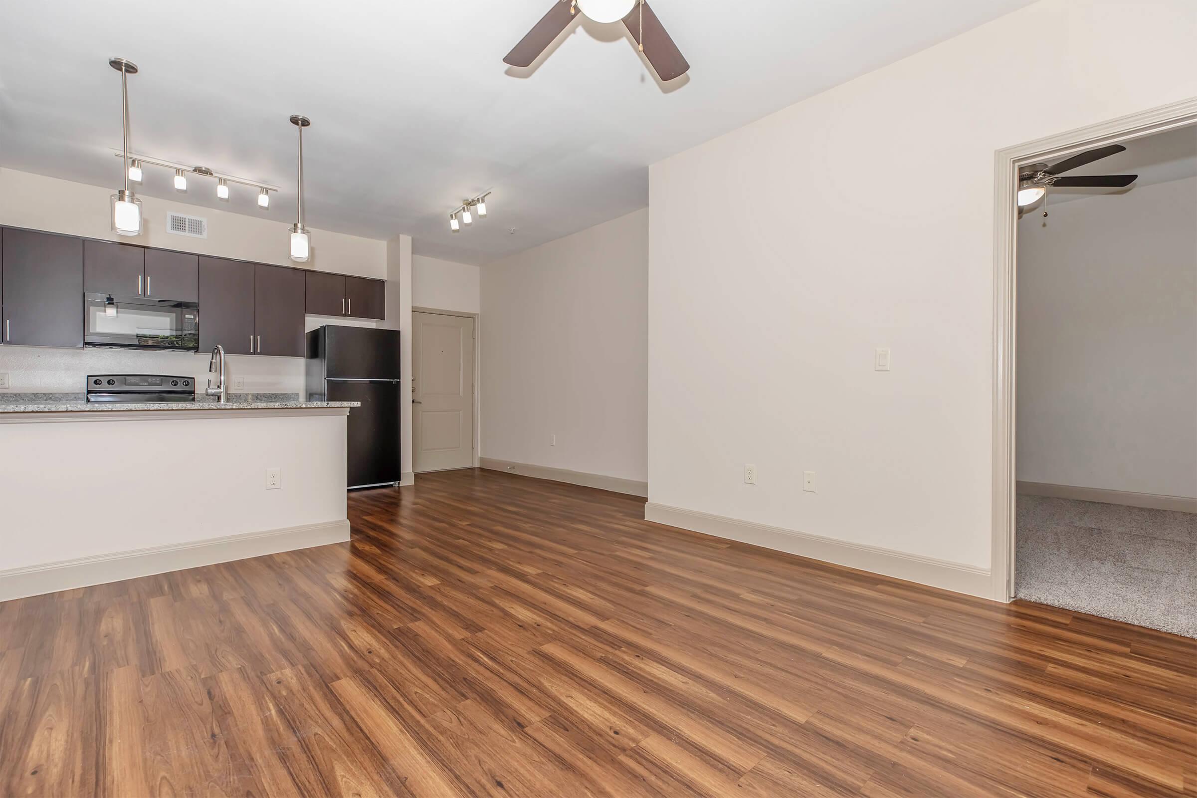 Spacious living area with hardwood flooring, featuring a kitchen bar with dark cabinetry and stainless steel appliances. Bright electric lights hang from the ceiling, and there's an open doorway leading to another room.
