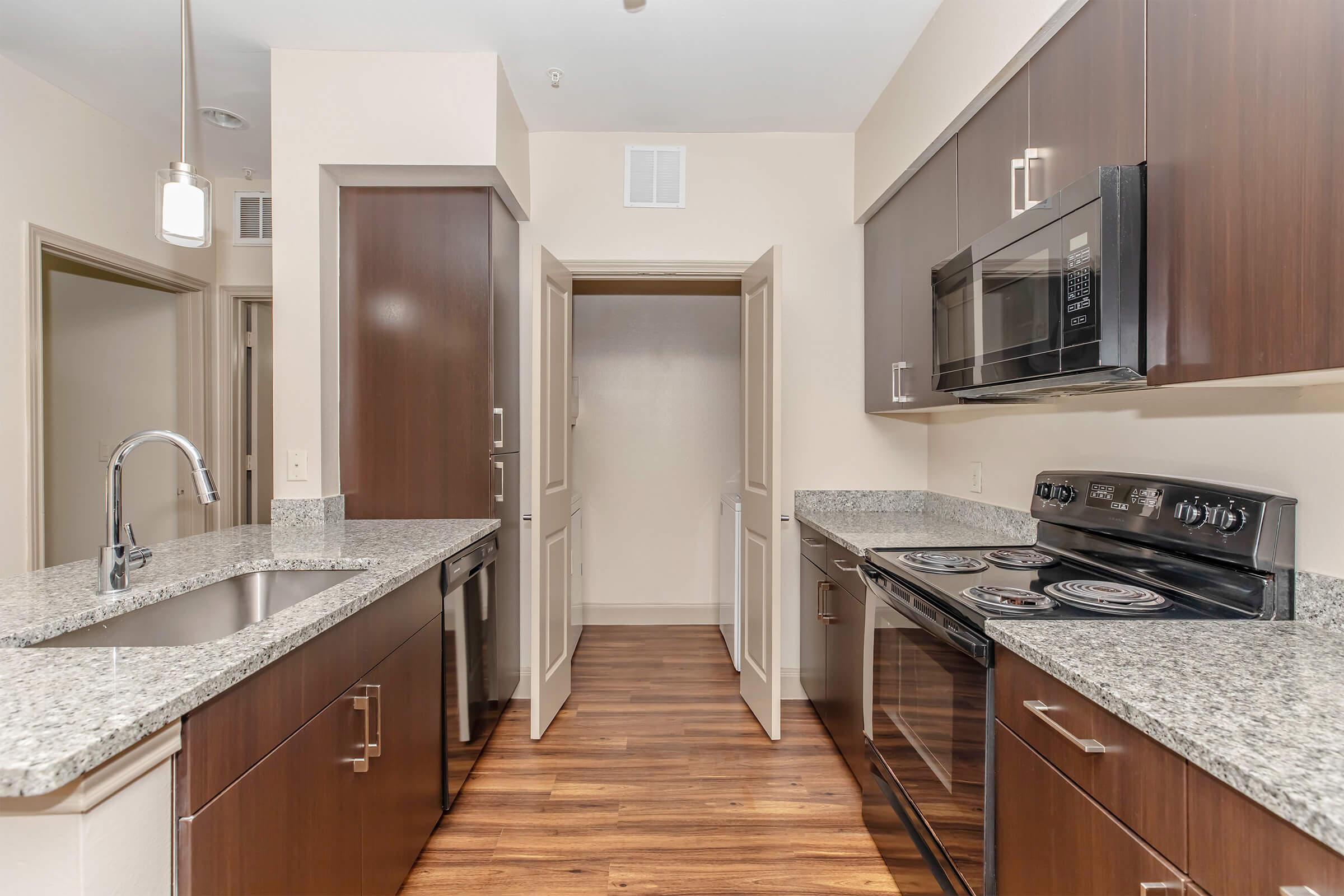 Modern kitchen featuring dark wood cabinetry, stainless steel appliances, and a granite countertop. The layout includes an island with a sink, an electric stove, and a microwave. A pantry door is visible, and the flooring is hardwood, giving the space a warm and inviting feel.