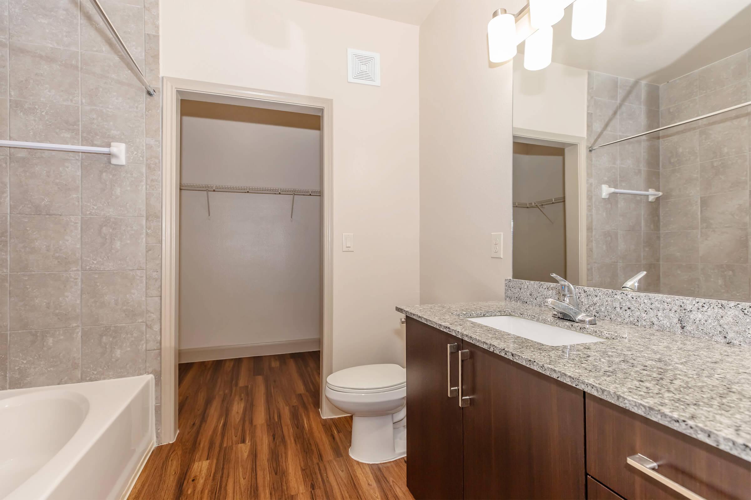 A modern bathroom featuring a granite countertop with a sink, a mirror above, a toilet, and a bathtub. The walls are tiled in light gray, and there is a wooden floor. A spacious closet is visible in the background, and a light fixture hangs from the ceiling.