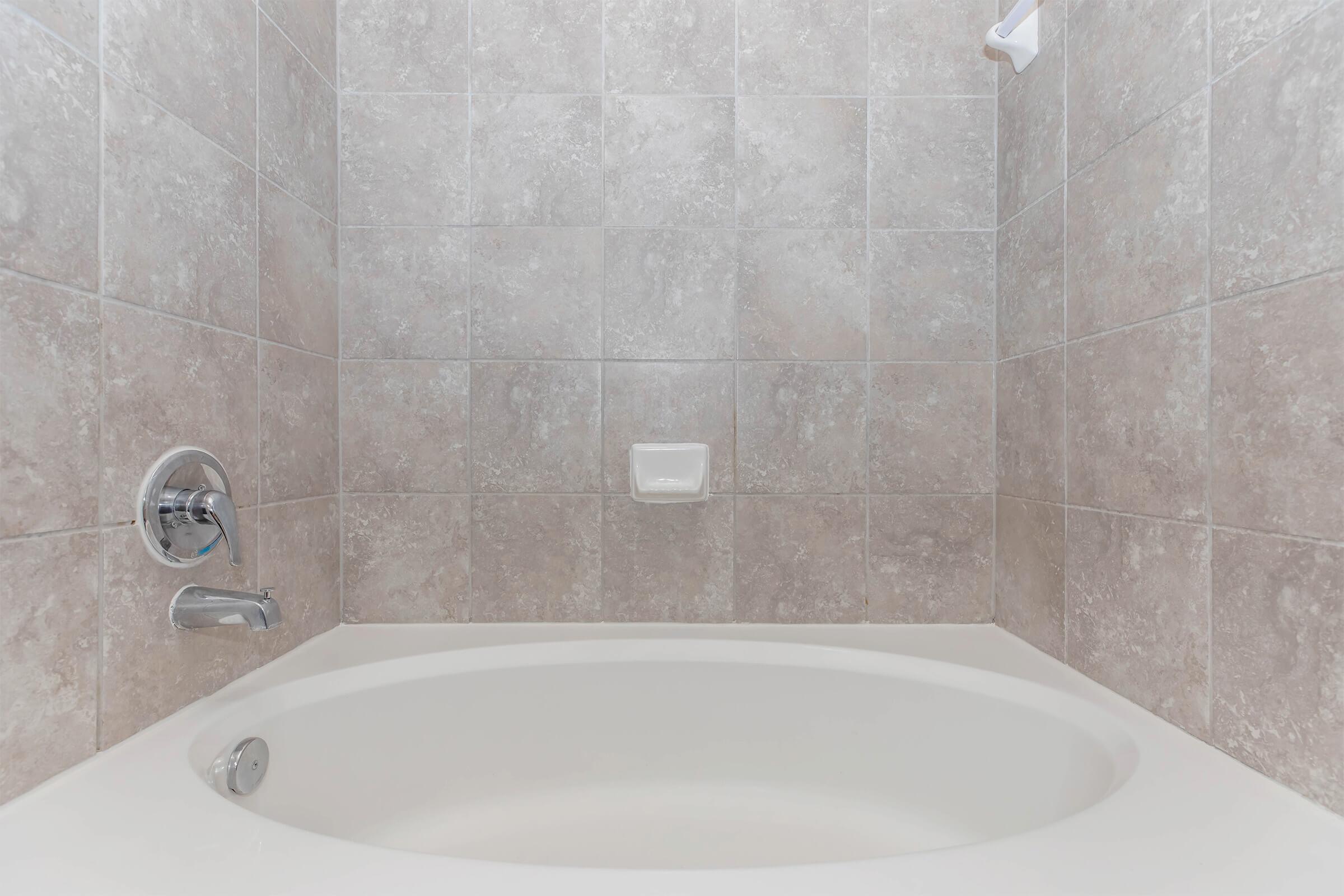 A spacious bathtub with a smooth, curved edge, set against a backdrop of light gray textured wall tiles. The bath faucet and a showerhead are visible, along with a soap dish mounted on the wall. The overall atmosphere suggests a clean and modern bathroom design.