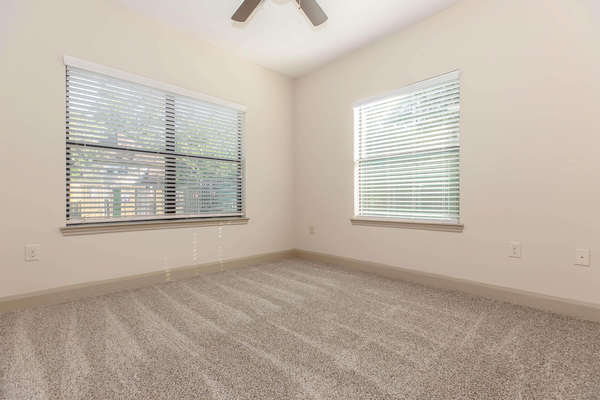 A well-lit empty room featuring light-colored walls and two windows with blinds. The floor is covered in soft beige carpet, and a ceiling fan is visible. The space is uncluttered, creating a feeling of openness and simplicity. Natural light streams in, enhancing the room's airy atmosphere.