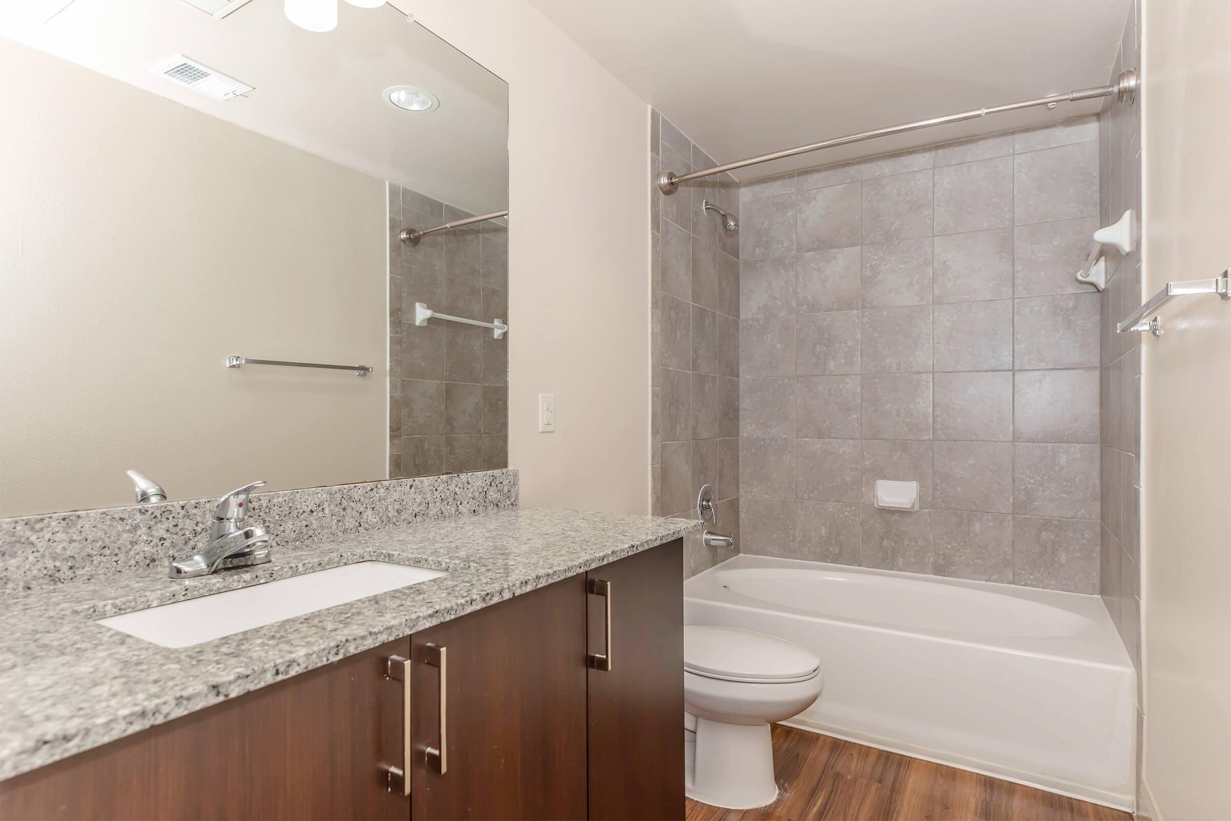 A modern bathroom featuring a bathtub with a tiled surround, a granite countertop sink area with a mirror above, and wooden flooring. The walls are painted in a neutral color, and there is a towel bar mounted on the wall. The overall design is sleek and contemporary.