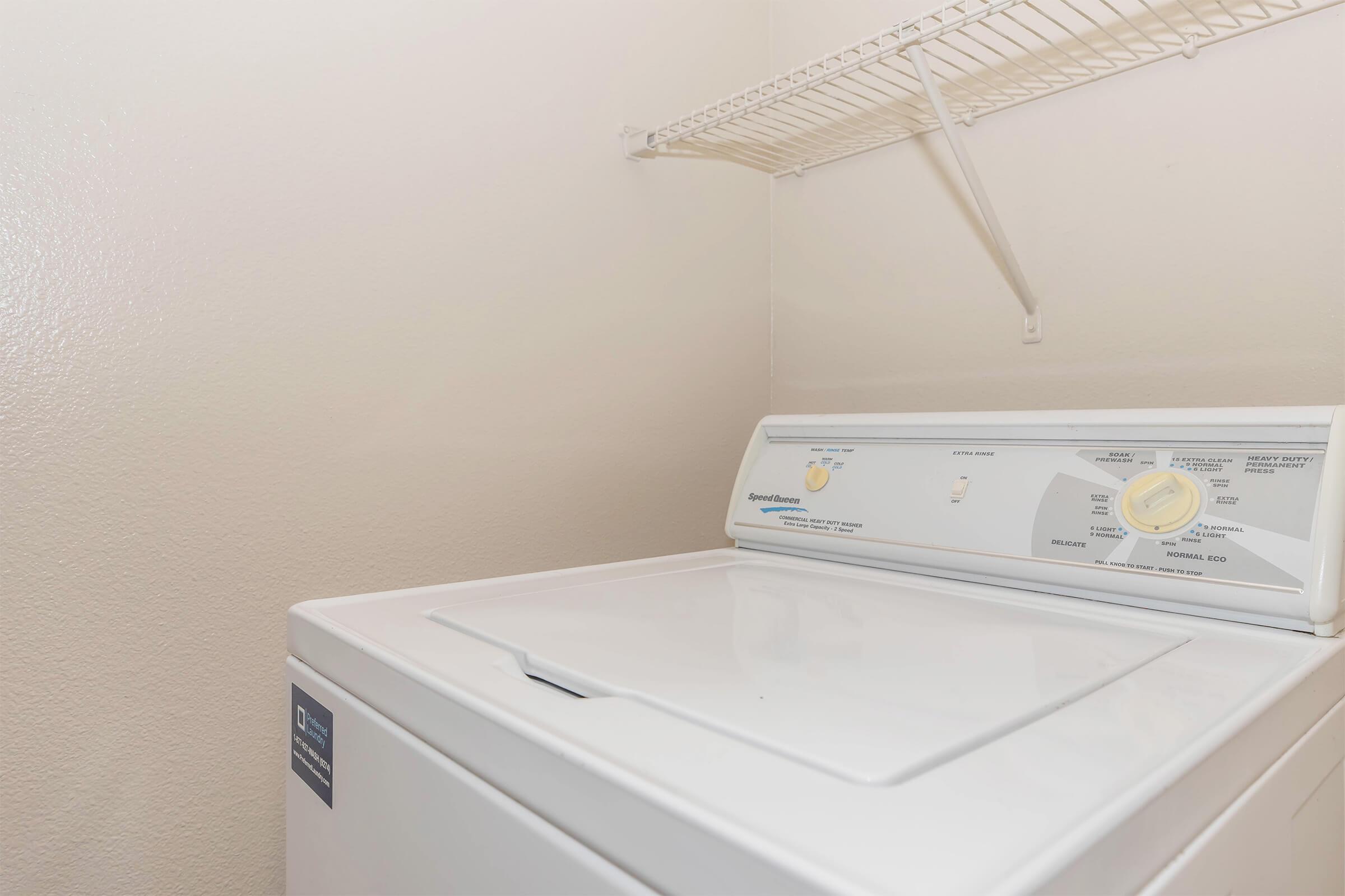 A white top-loading washing machine positioned against a light-colored wall, with a wire shelf above it. The machine has control dials and labels for different wash settings. The overall setting appears to be a laundry room or utility space.