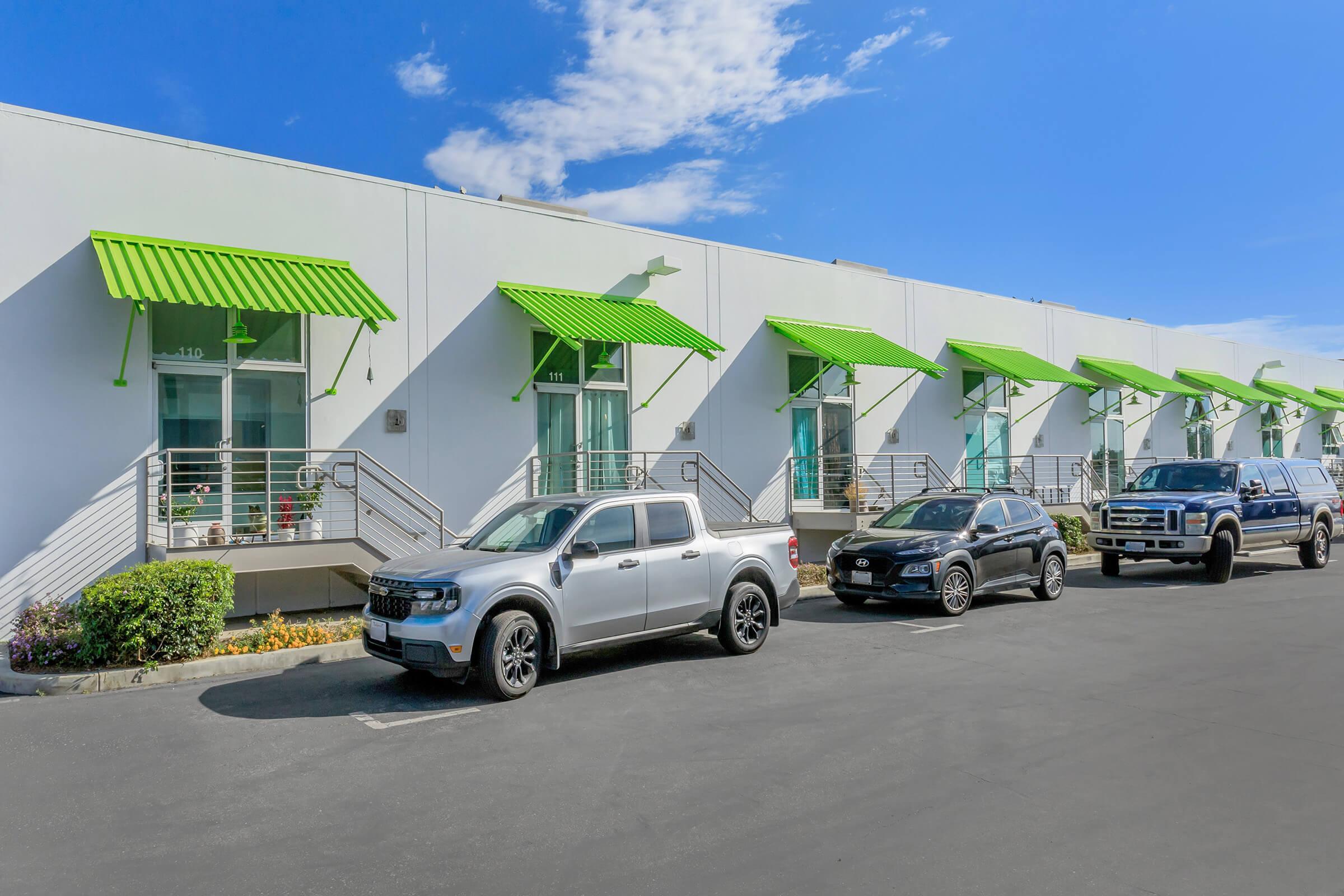 A row of commercial buildings with green awnings over the entrance doors. In the foreground, three parked vehicles—a silver pickup truck, a black sedan, and a dark SUV—are lined up along a paved parking area. The sky is clear with a few clouds.