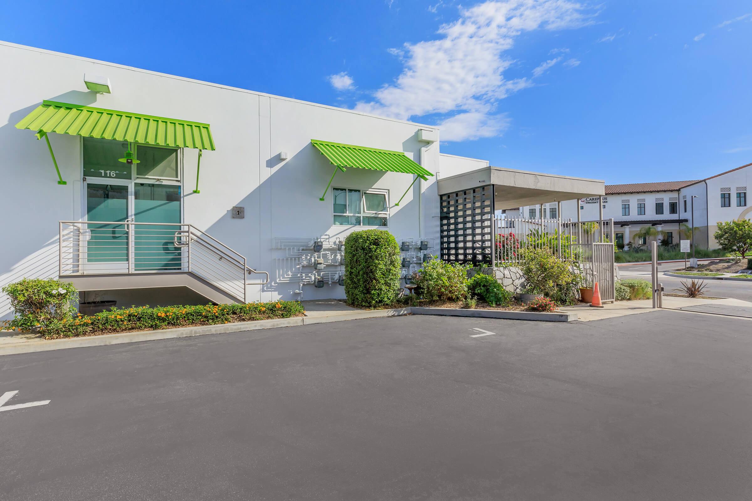 Modern commercial building with bright green awnings, a glass entrance, and landscaping featuring shrubs and flowers. A clear blue sky is visible above, and a portion of a parking lot is shown in the foreground. The scene conveys a fresh and inviting atmosphere.