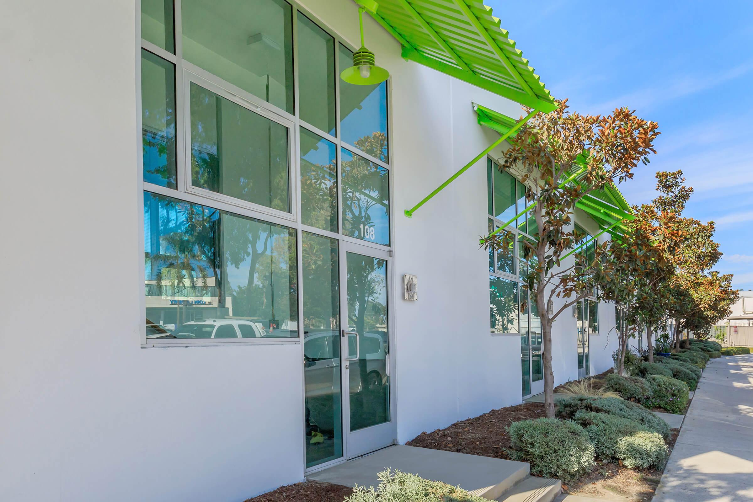 A modern building with large windows and a bright green overhang. The entrance features a glass door, and there are neatly trimmed shrubs and trees alongside a walkway. The sky is clear with a few clouds, creating a bright and inviting atmosphere.
