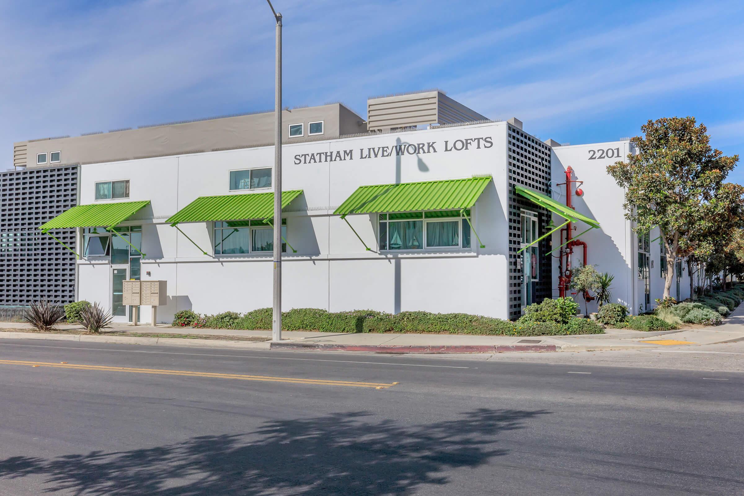 Exterior view of Statham Live/Work Lofts, a modern building featuring white walls, green awnings, and large windows. Vibrant landscaping with shrubs and trees is visible in front. The street runs parallel to the building, and a clear blue sky is overhead. The building number is 2201.
