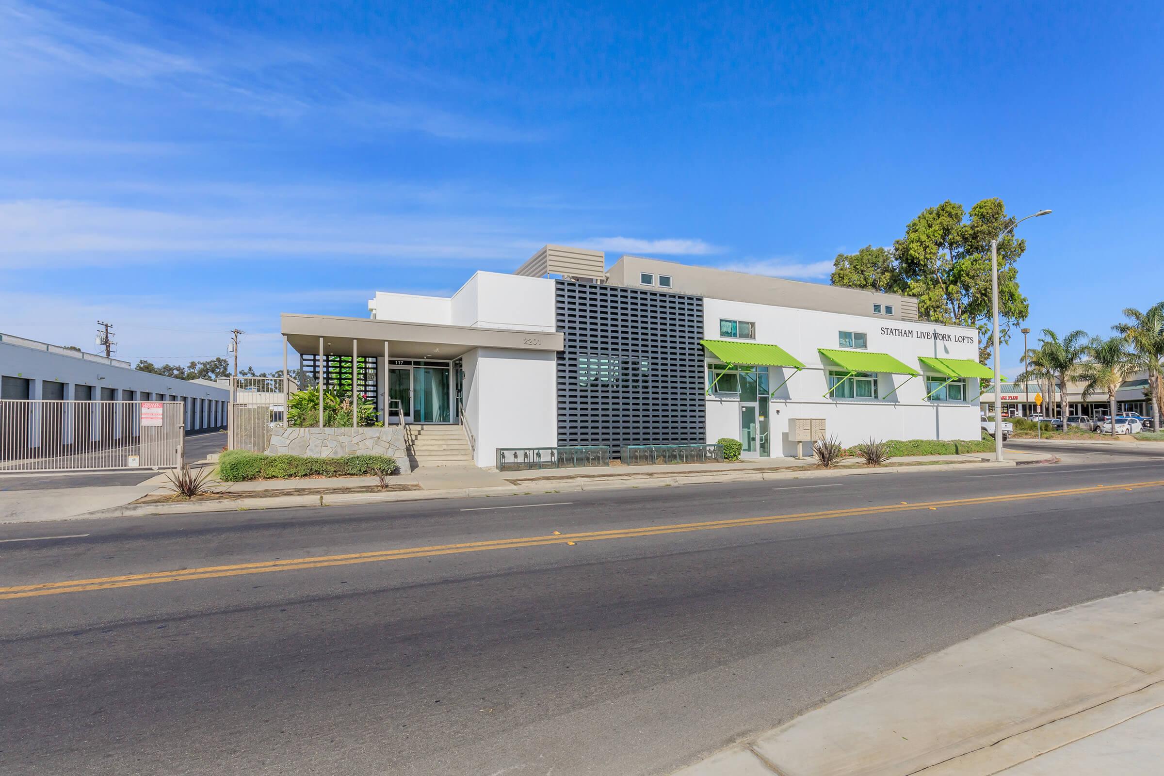 A modern commercial building with large windows and green awnings, situated on a street corner. The structure features a light-colored facade and is surrounded by palm trees and landscaping. Clear blue sky above enhances the contemporary look of the architecture.