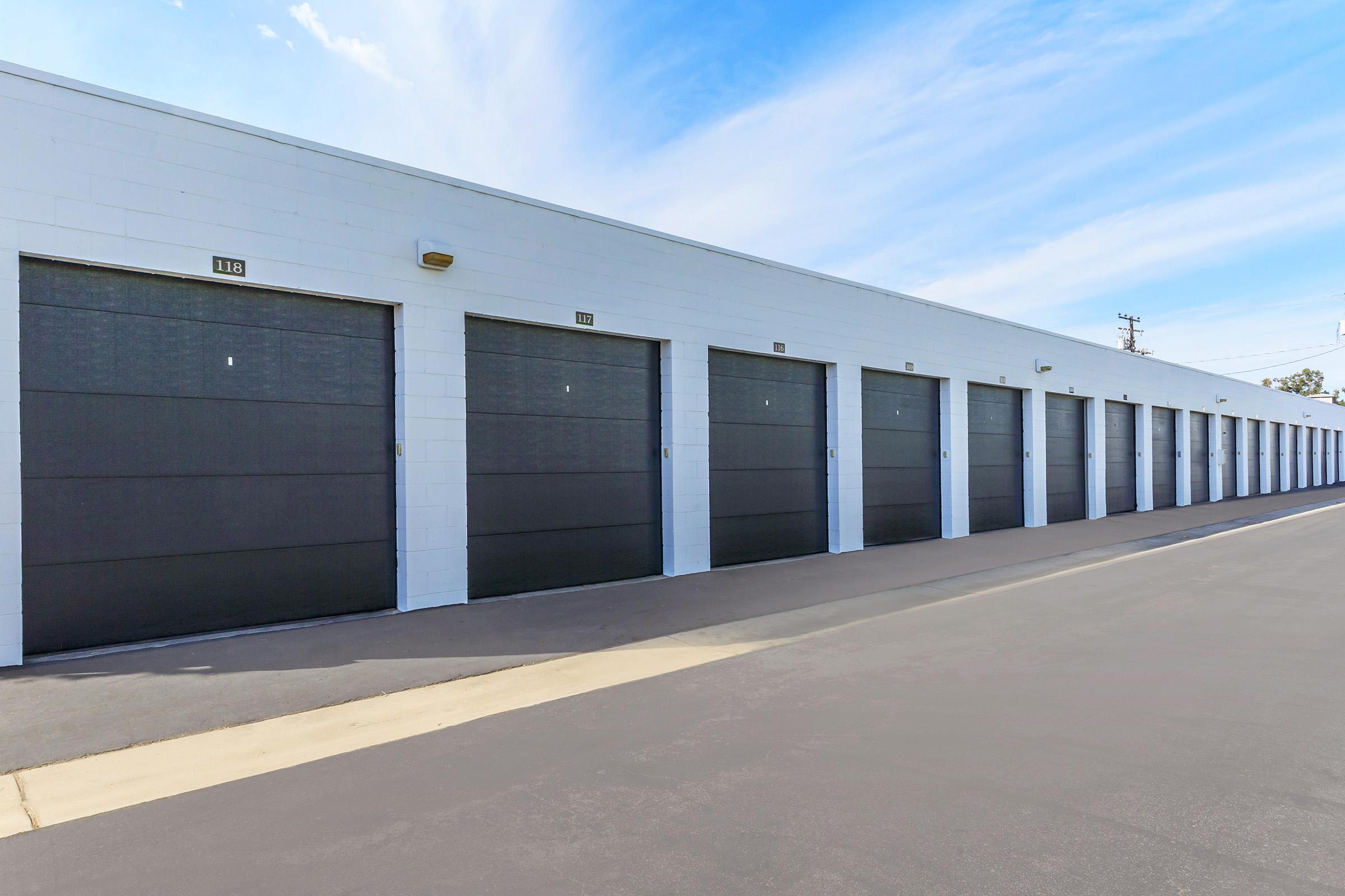 A row of storage units with closed metal doors, set against a clear blue sky. The units are neatly aligned on a paved surface, showcasing a well-maintained facility.