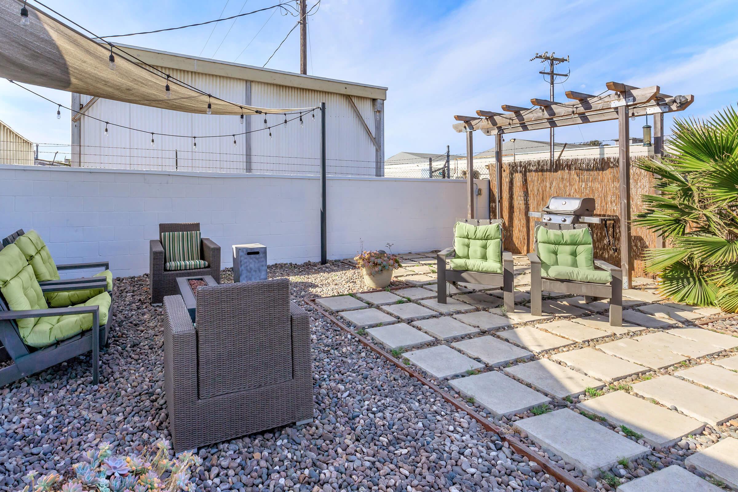A cozy outdoor patio featuring green cushioned chairs and a shaded seating area under a pergola. The space is decorated with gravel and stone pavers, surrounded by wooden fencing and palm plants, creating a relaxing environment for gatherings or quiet moments.