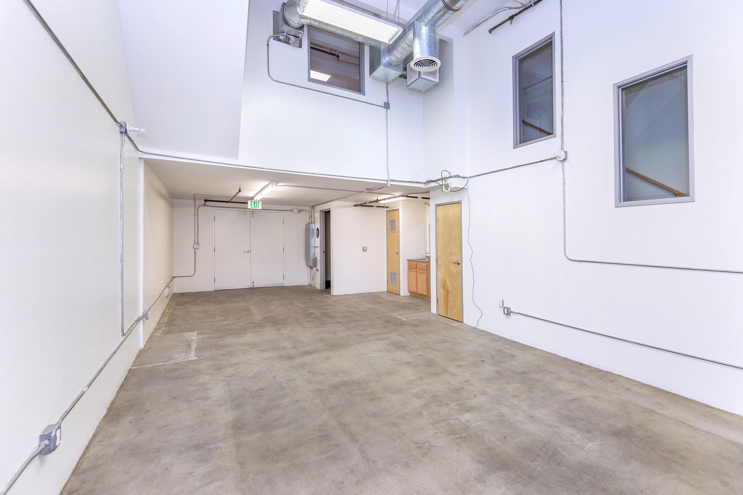 An empty indoor space with concrete flooring, white walls, and exposed pipes. There are two small windows and a door on one side, with additional doors on the opposite wall. The area is well-lit and has an industrial feel, suitable for various uses such as storage or a workshop.