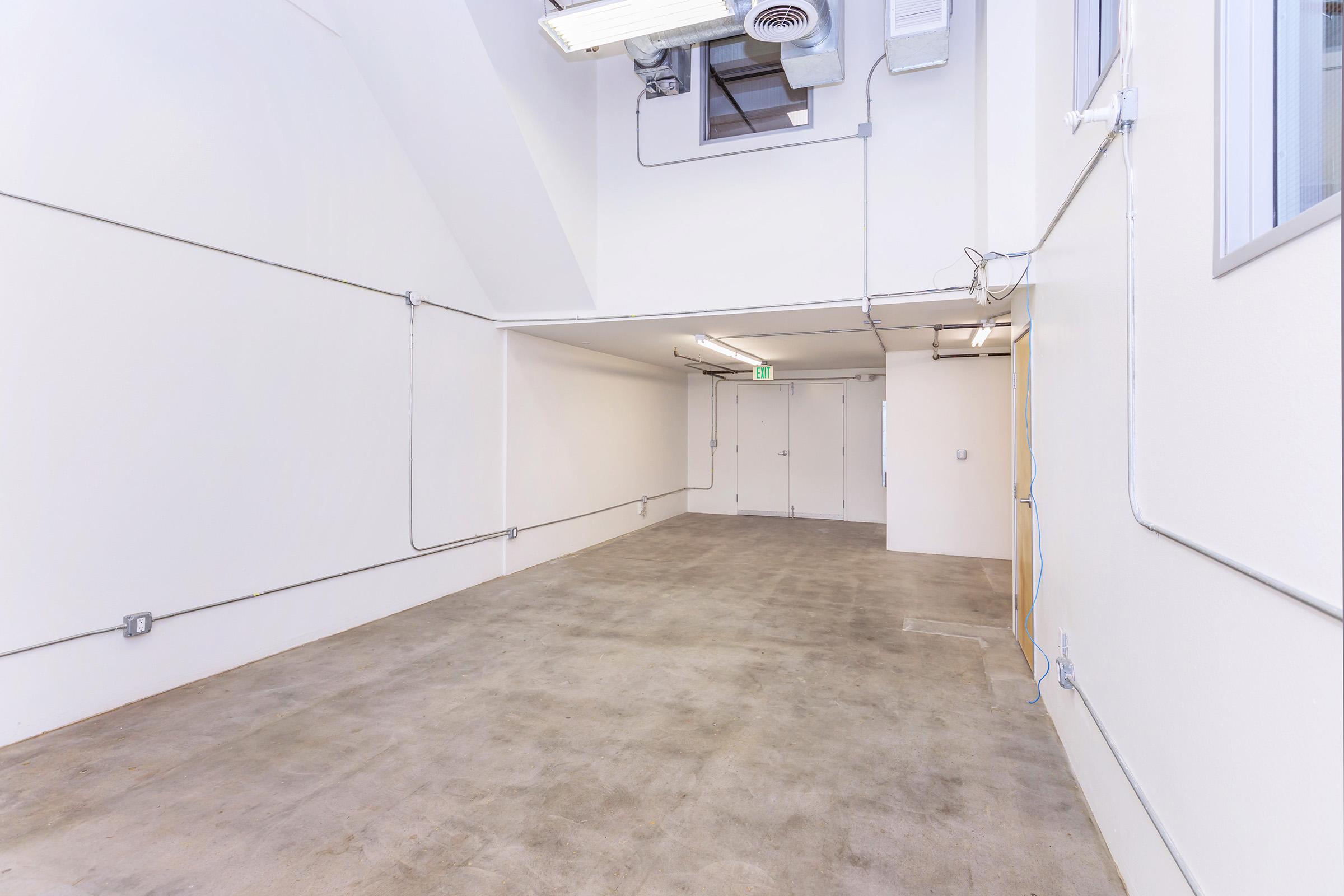 A spacious, empty room with concrete flooring and white walls. Electrical wires are exposed along the walls and ceiling. There are double doors at the far end, and the room is well-lit with fluorescent lighting overhead, creating a bright, open atmosphere.