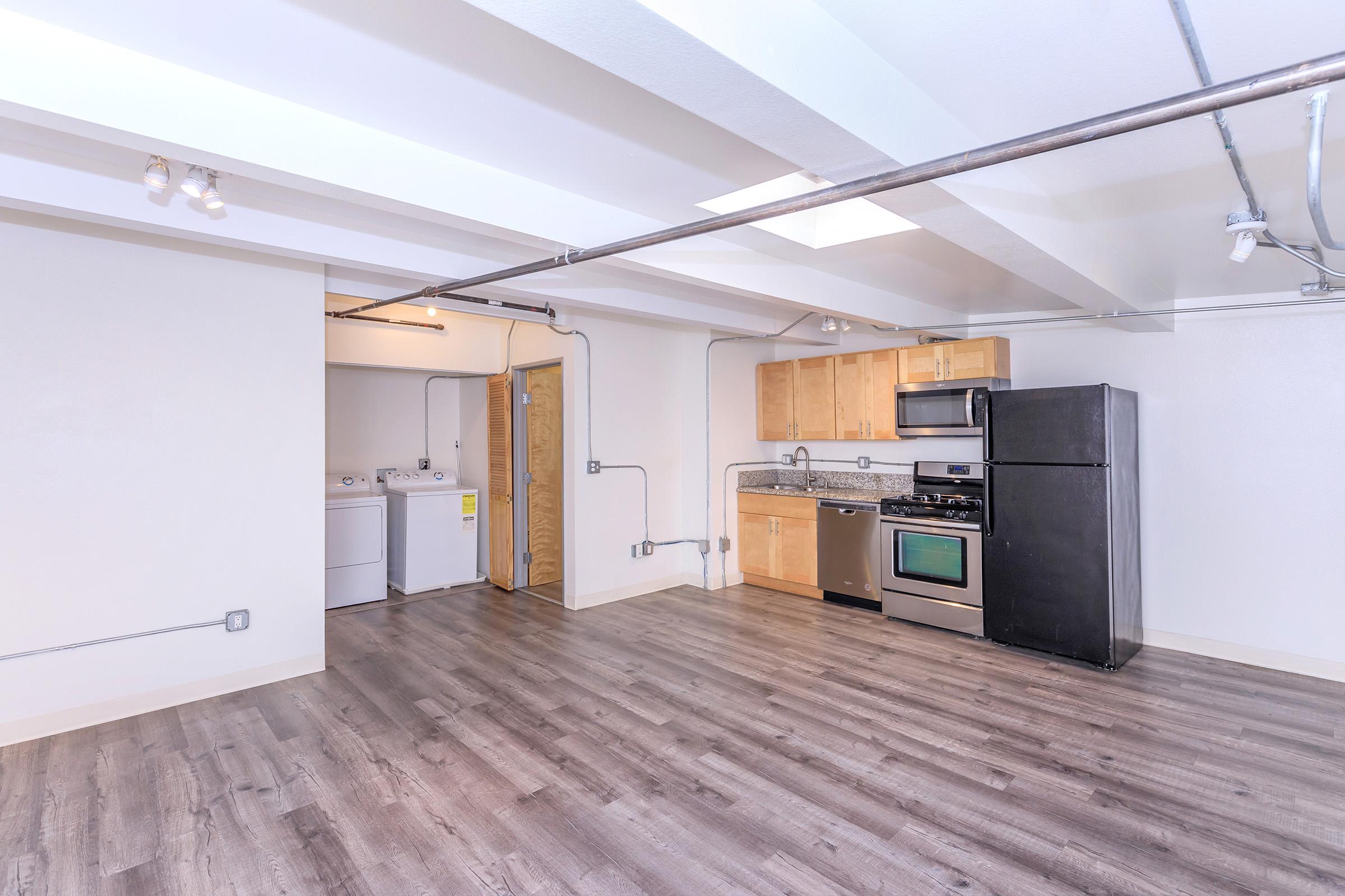 Modern open-concept kitchen and living area featuring stainless steel appliances including a stove and refrigerator, wooden cabinets, and a laundry area with a washer and dryer. The room has a hardwood floor and exposed ductwork, with natural light coming from a skylight.