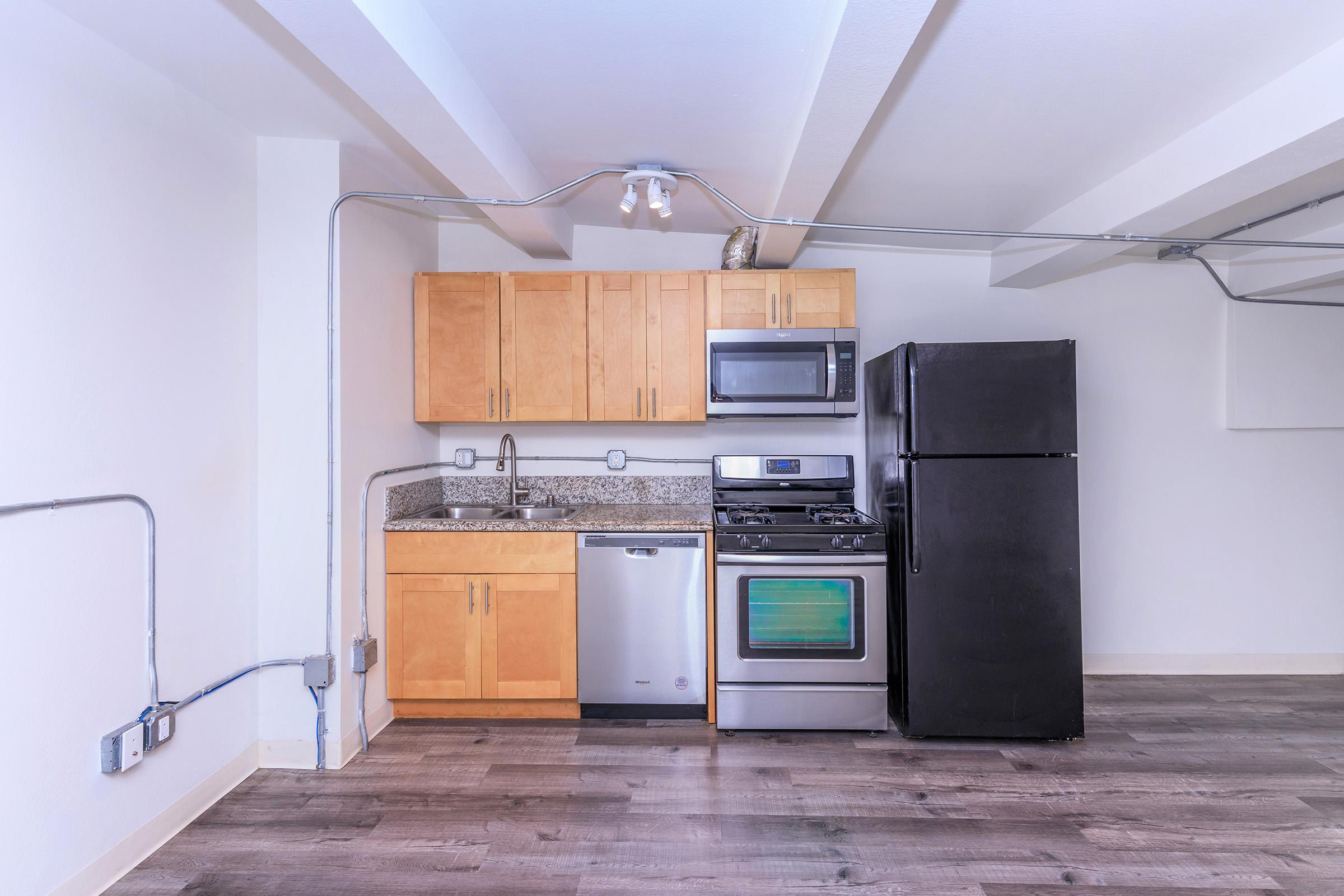 Modern kitchen in an apartment featuring wooden cabinets, granite countertops, stainless steel appliances including a microwave, gas stove, dishwasher, and a black refrigerator. Exposed beams and pipes add an industrial touch to the design.