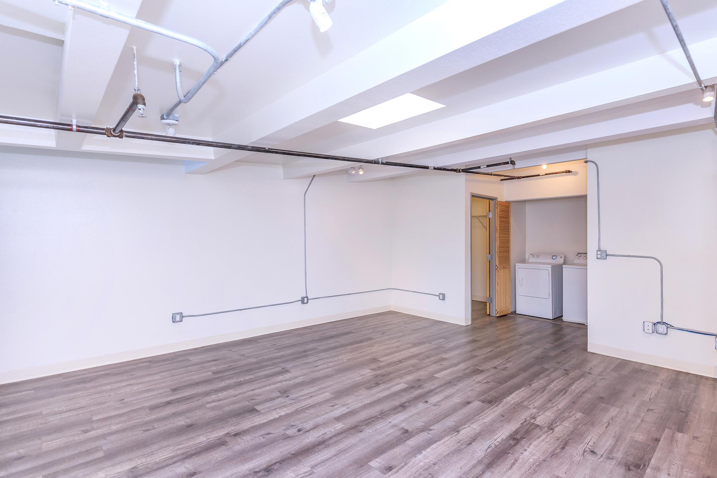 A spacious, empty room showcasing light-colored walls and a wood-like floor. Exposed pipes run across the ceiling, and a laundry area with a washer and dryer is visible in the back corner. Natural light enters through a ceiling window, creating a bright and open atmosphere.