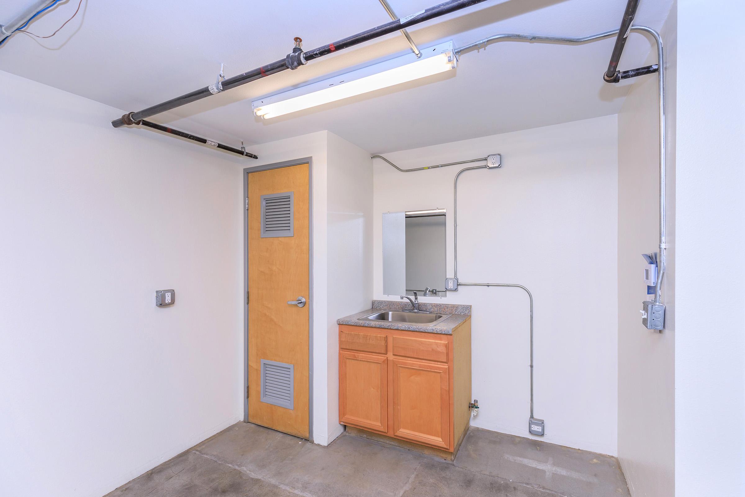 A simple, uncluttered utility room featuring a small wooden sink cabinet with a countertop, a mirror above it, and exposed pipes along the ceiling and walls. A wooden door is visible, and the floor is concrete. The lighting is bright and fluorescent, creating a functional, minimal space.